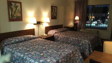 A hotel room with two beds, each with floral bedding, bedside lamps, and a nightstand with a phone. There is a window with a view of the city at night.