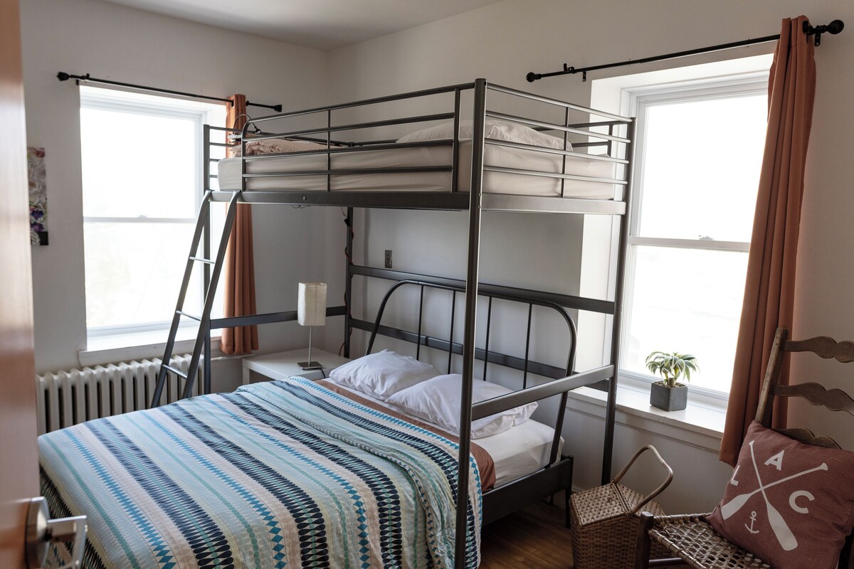 A bunk bed room with a window, a bedside table, a lamp, and a potted plant.