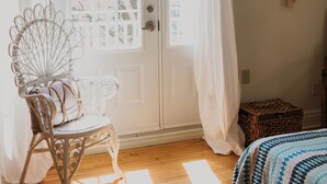 A room with a wooden floor, a white door with curtains, a wicker chair, a bed with a patterned bedspread, and a macrame wall hanging. - Auberge Jeunesse Magog-Orford (Hostel) (Magog)
