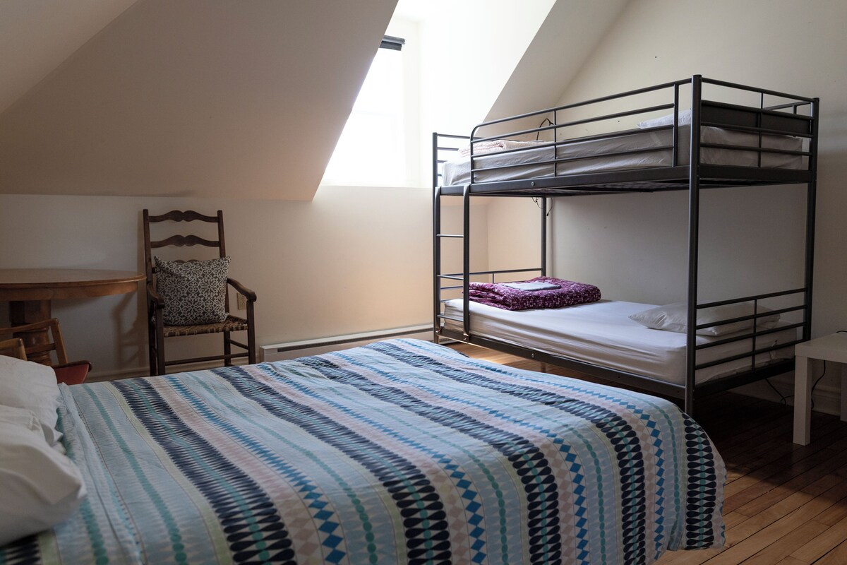 A dormitory room with bunk beds, a desk, a chair, and a window providing natural light.