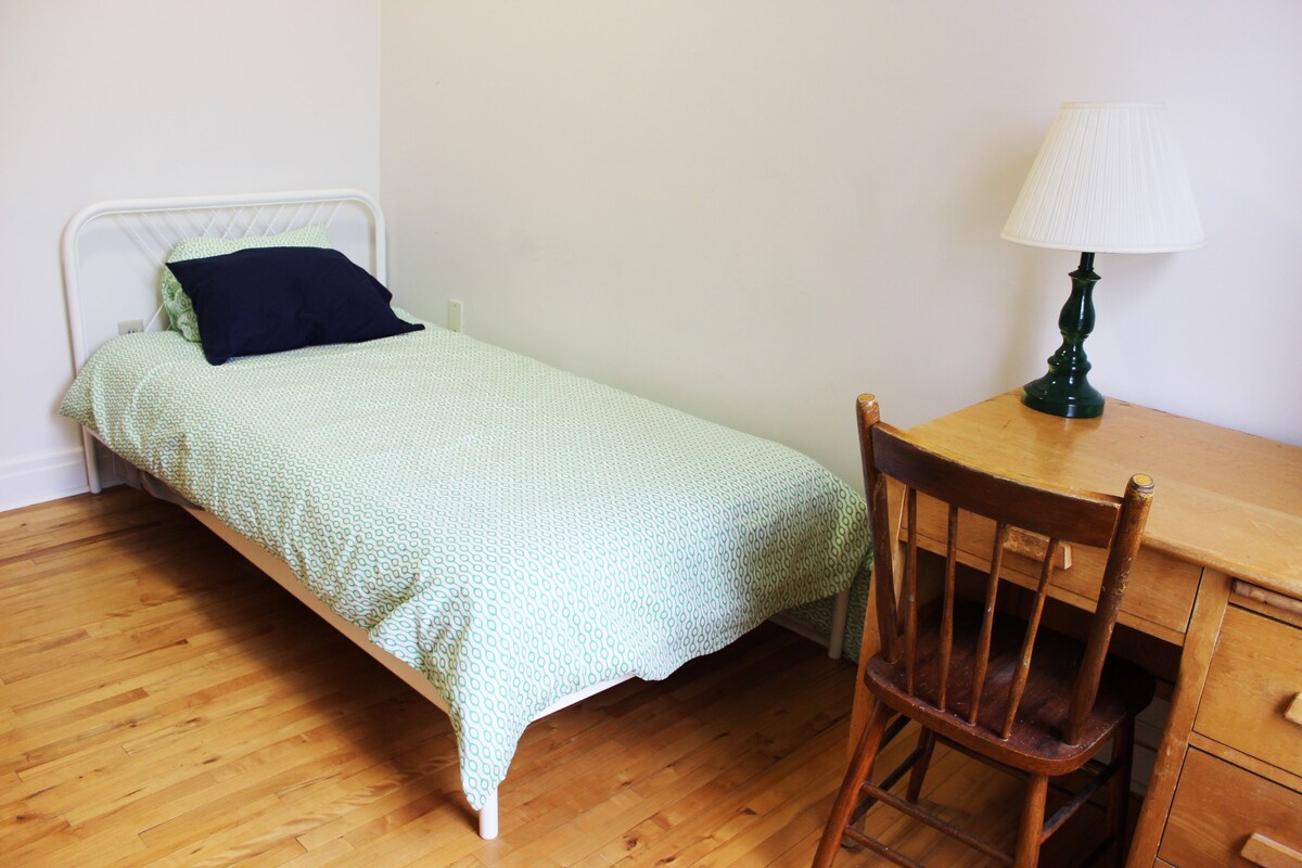 A small, single-bed room with a wooden floor, a green patterned bedspread, a dark pillow, a wooden chair, and a green table lamp.
