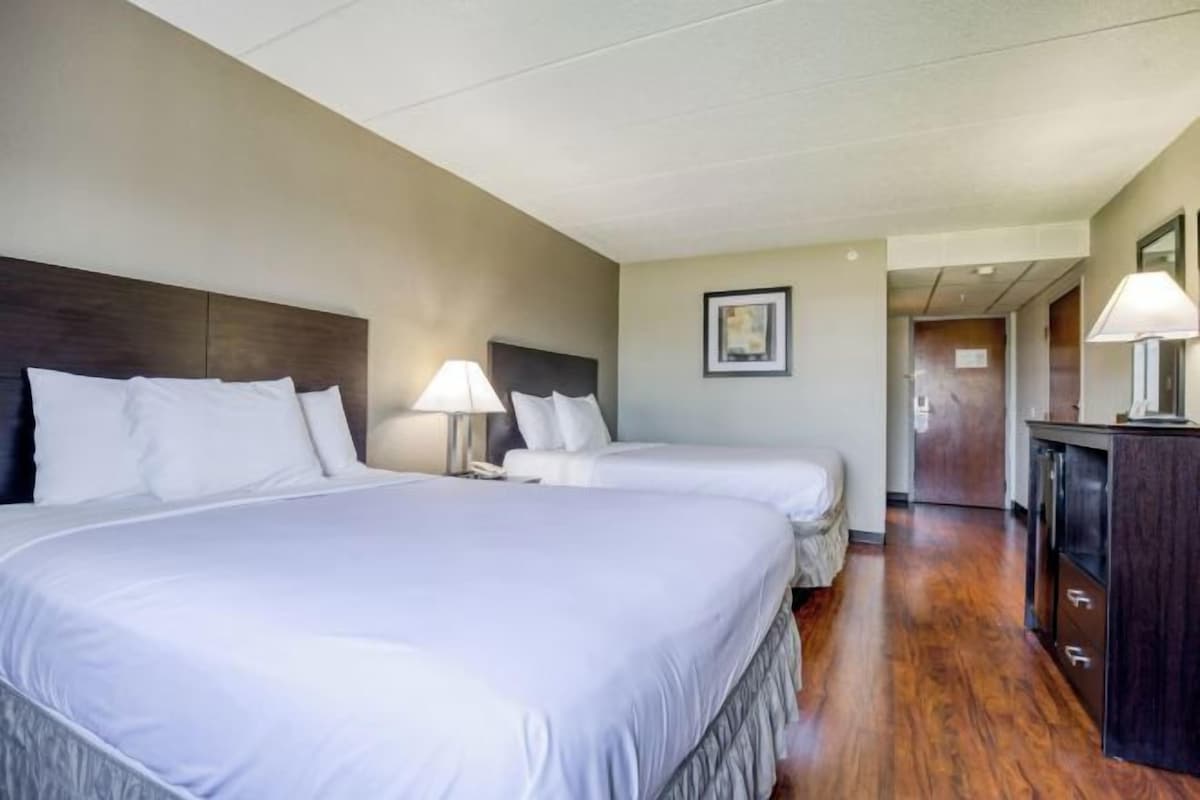 A hotel room with two beds, a wooden floor, a nightstand with a lamp, and a door leading to another room.