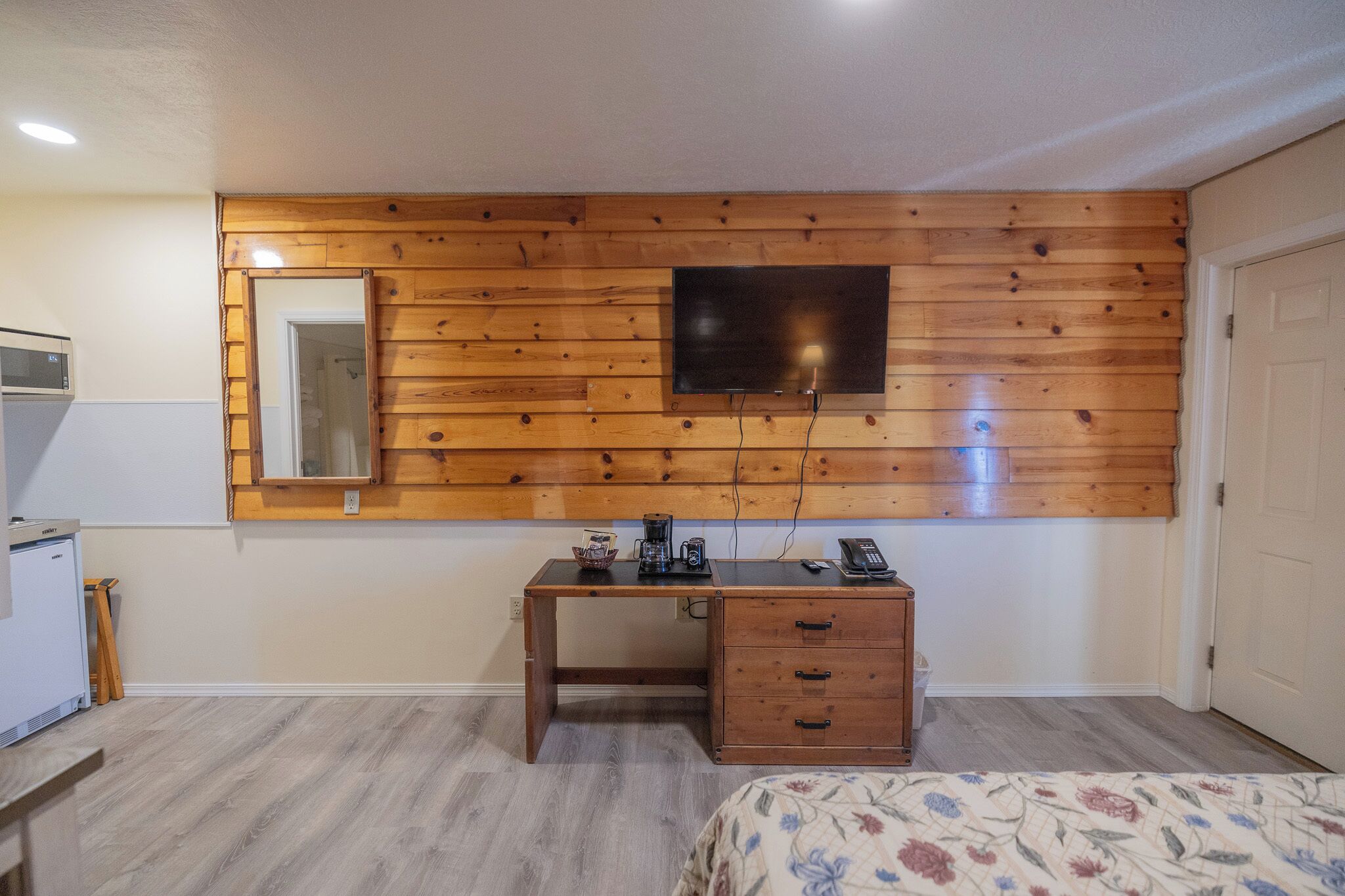 Une chambre avec un panneau mural en bois, un lit, un bureau avec des tiroirs, un miroir et un téléviseur fixé au mur.