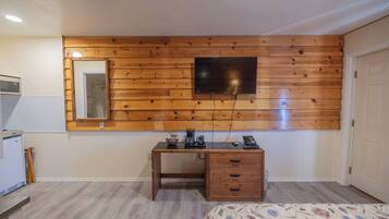 A bedroom with a wooden wall panel, a bed, a desk with drawers, a mirror, and a television mounted on the wall.