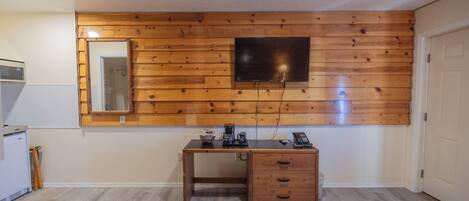 Une chambre avec un panneau mural en bois, un lit, un bureau avec des tiroirs, un miroir et un téléviseur fixé au mur.