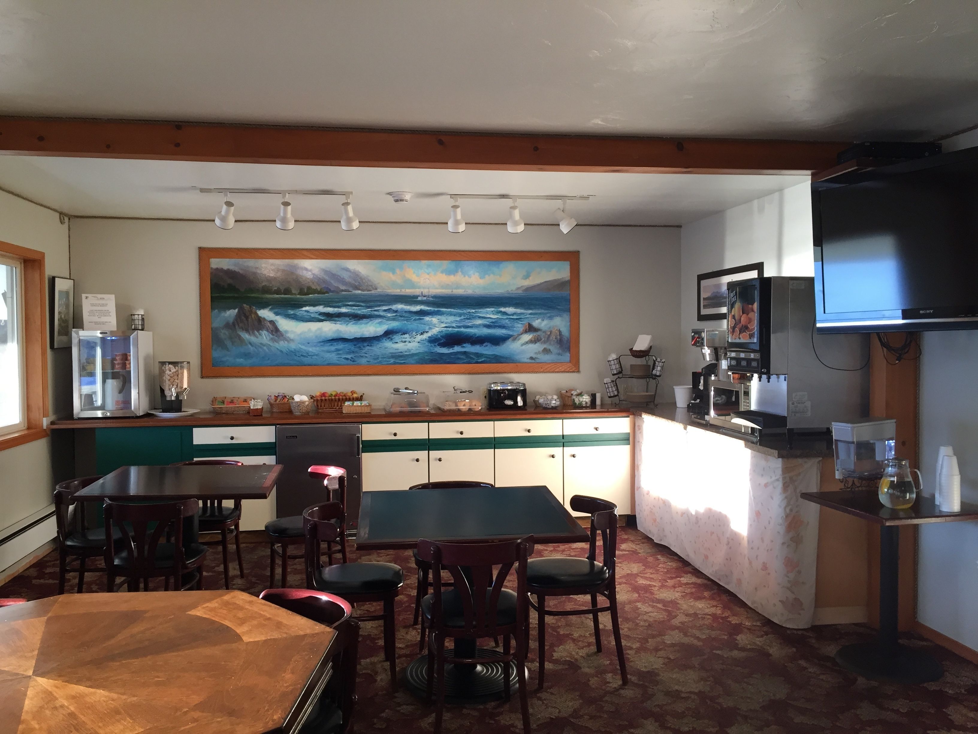 A restaurant with a large ocean painting, wooden ceiling beams, a coffee machine, and a TV.