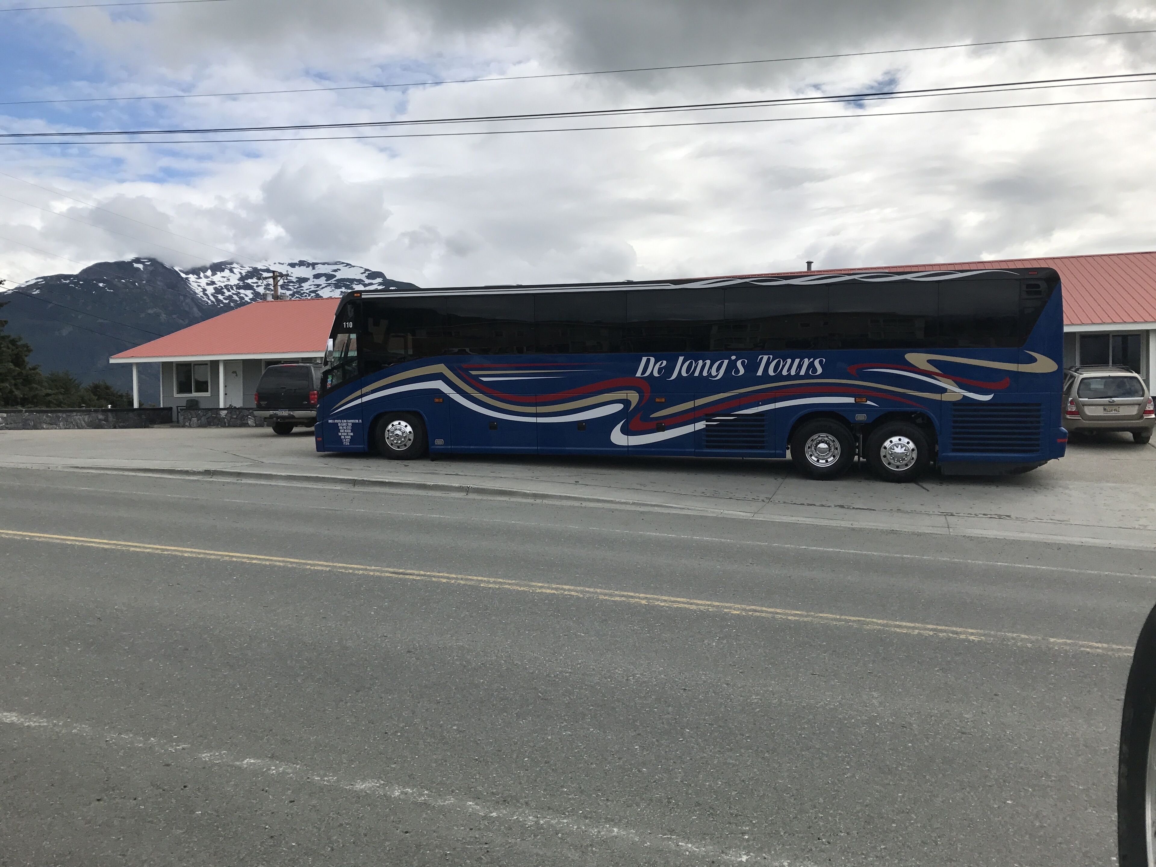 A blue tour bus with "De Jong's Tours" branding parked on a road with a building and mountains in the background.