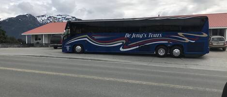 A blue tour bus with "De Jong's Tours" branding parked on a road with a building and mountains in the background.