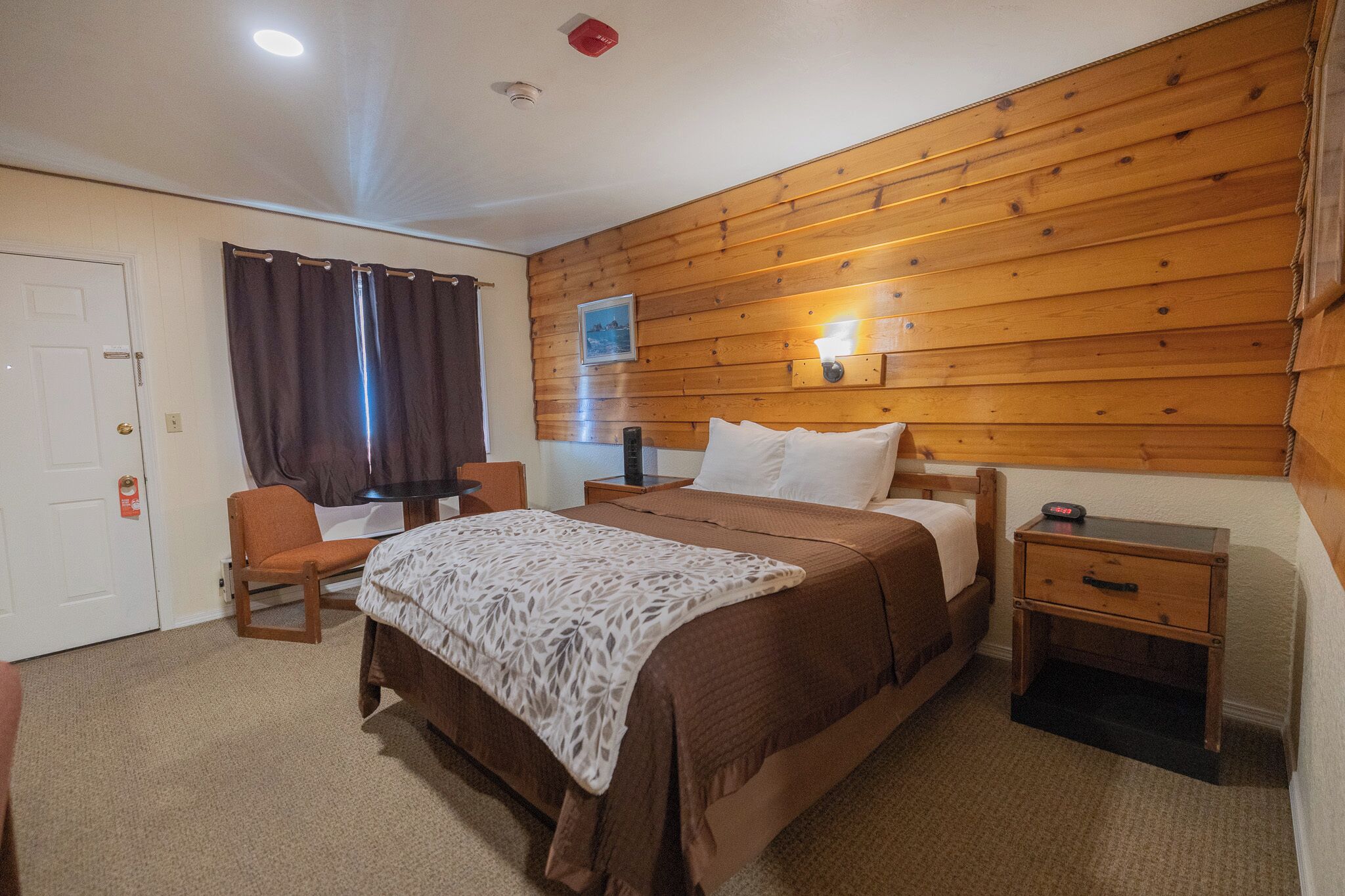 A bedroom with a wooden wall, a bed with a white and brown bedspread, a nightstand with a lamp, and a chair.