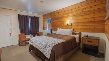 A bedroom with a wooden wall, a bed with a white and brown bedspread, a nightstand with a lamp, and a chair.