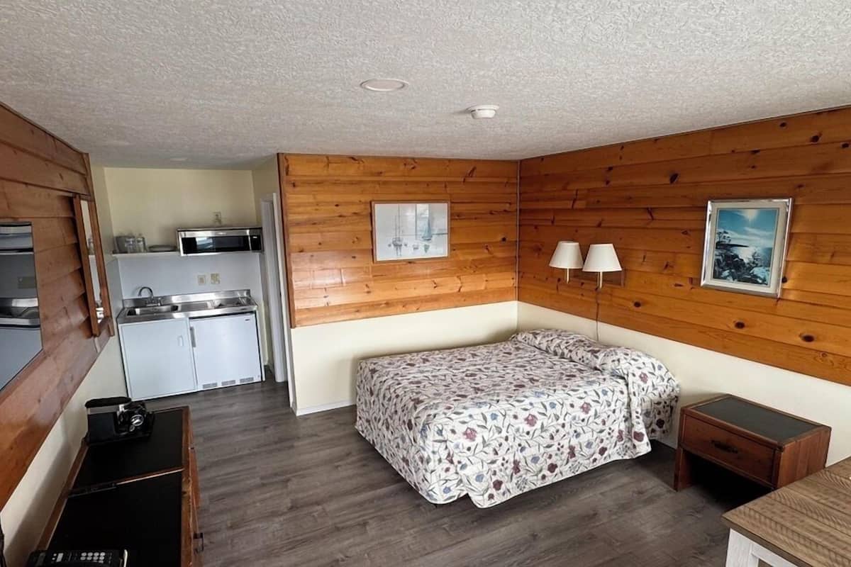 A compact room with a bed, a small table, a desk, a kitchenette, and wooden walls.