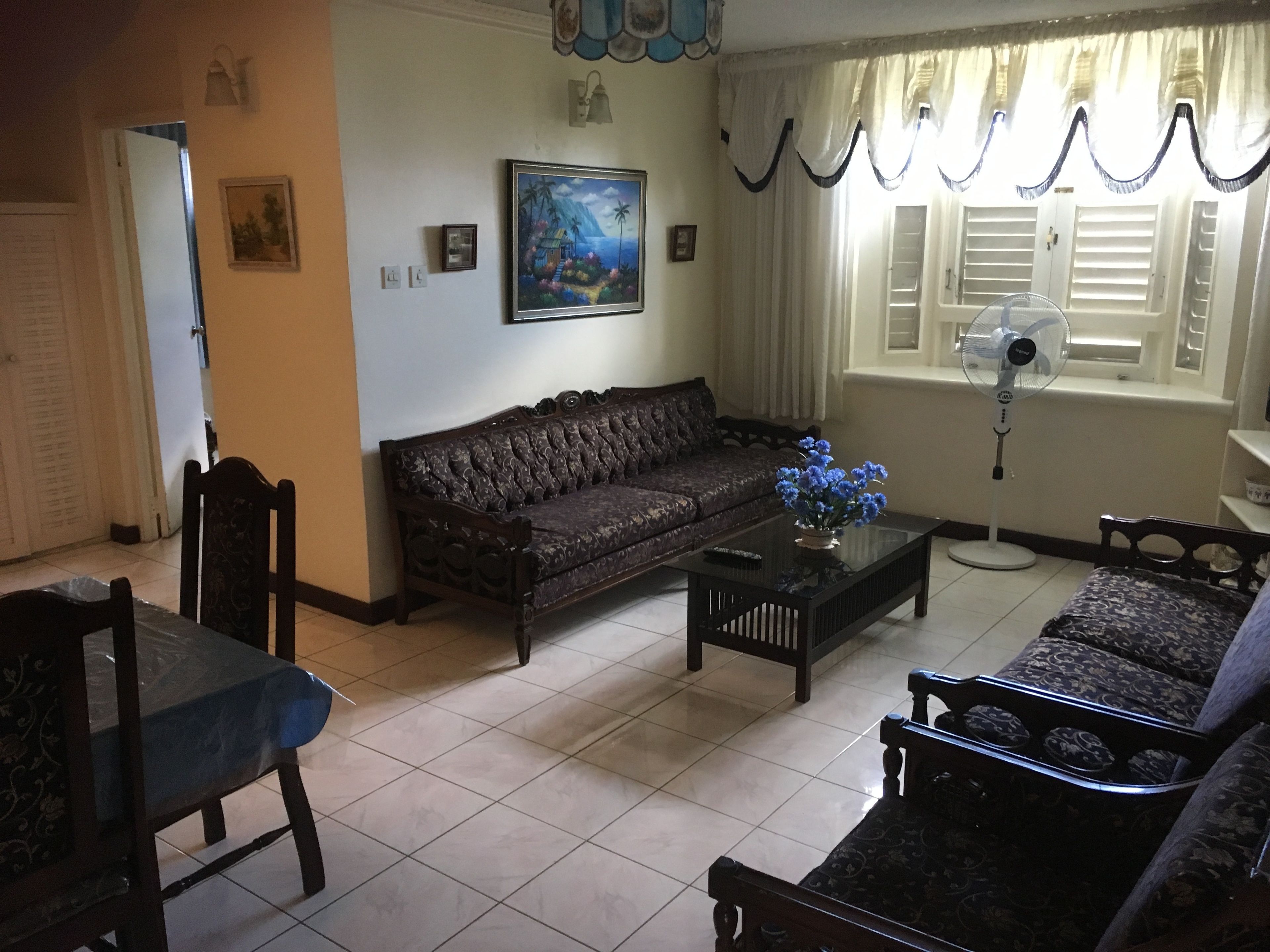 Ein Wohnzimmer mit einer Couch, einem Couchtisch mit Blumen, einem Ventilator und einem Fenster mit Vorhängen.