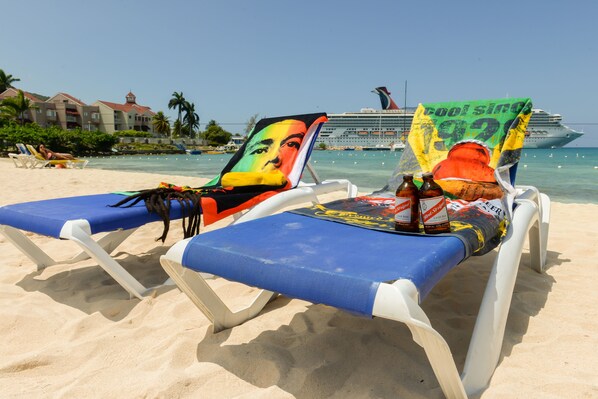 Two beach chairs with colorful artwork and a beverage cooler, a cruise ship in the background, and a hotel building with a red roof.