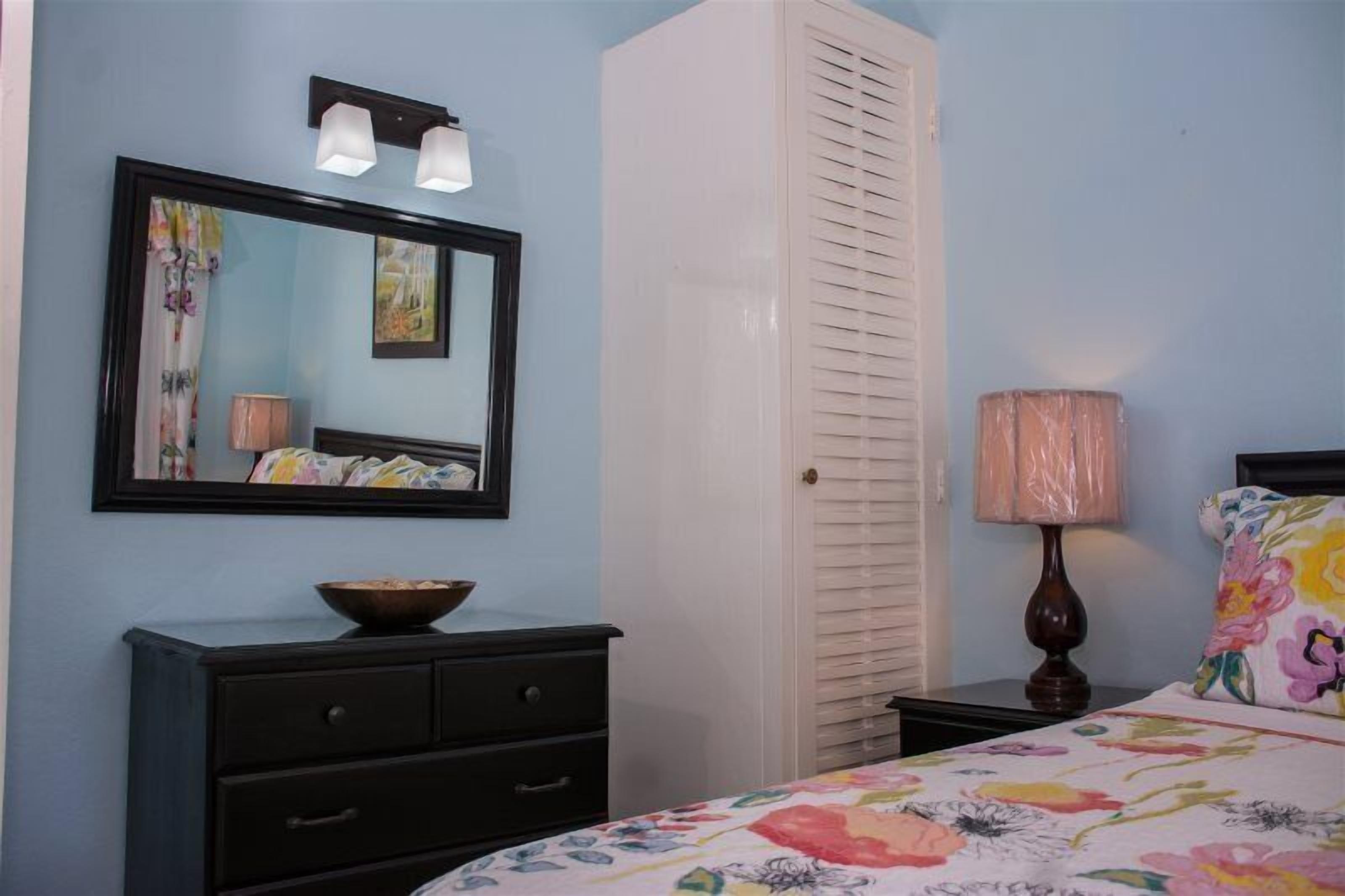 A bedroom with a bed, a dresser, a mirror, a window with blinds, and a nightstand with a lamp.