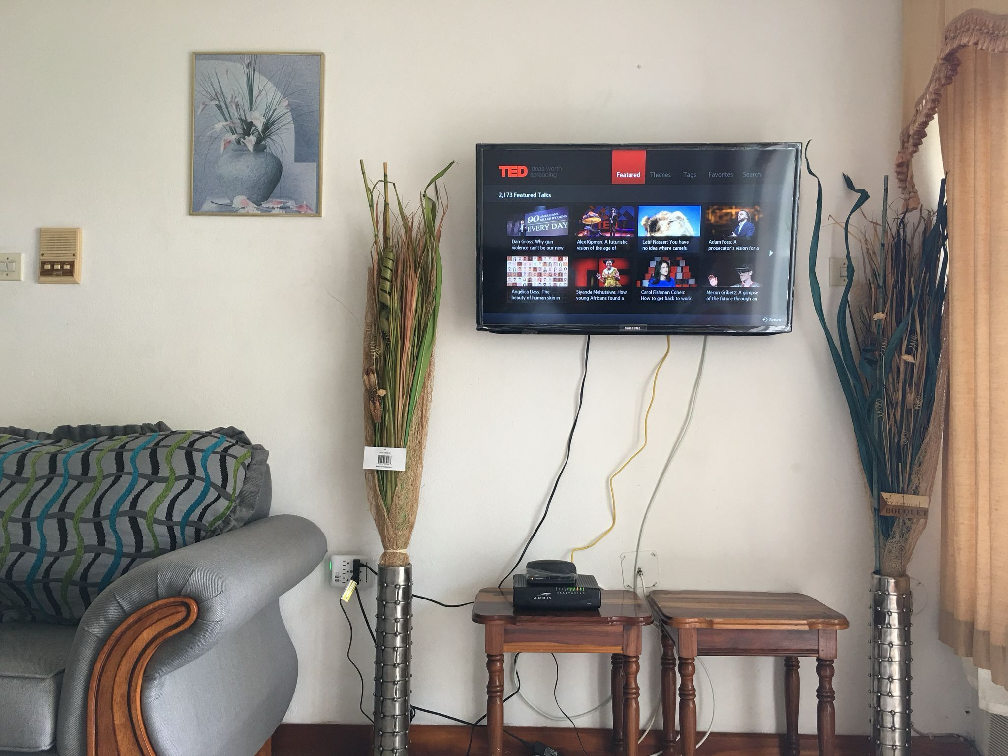 Een woonkamer met een grijze bank, een houten salontafel, een aan de muur bevestigde televisie waarop TED Talks worden vertoond, en een vaas met gedroogde planten.