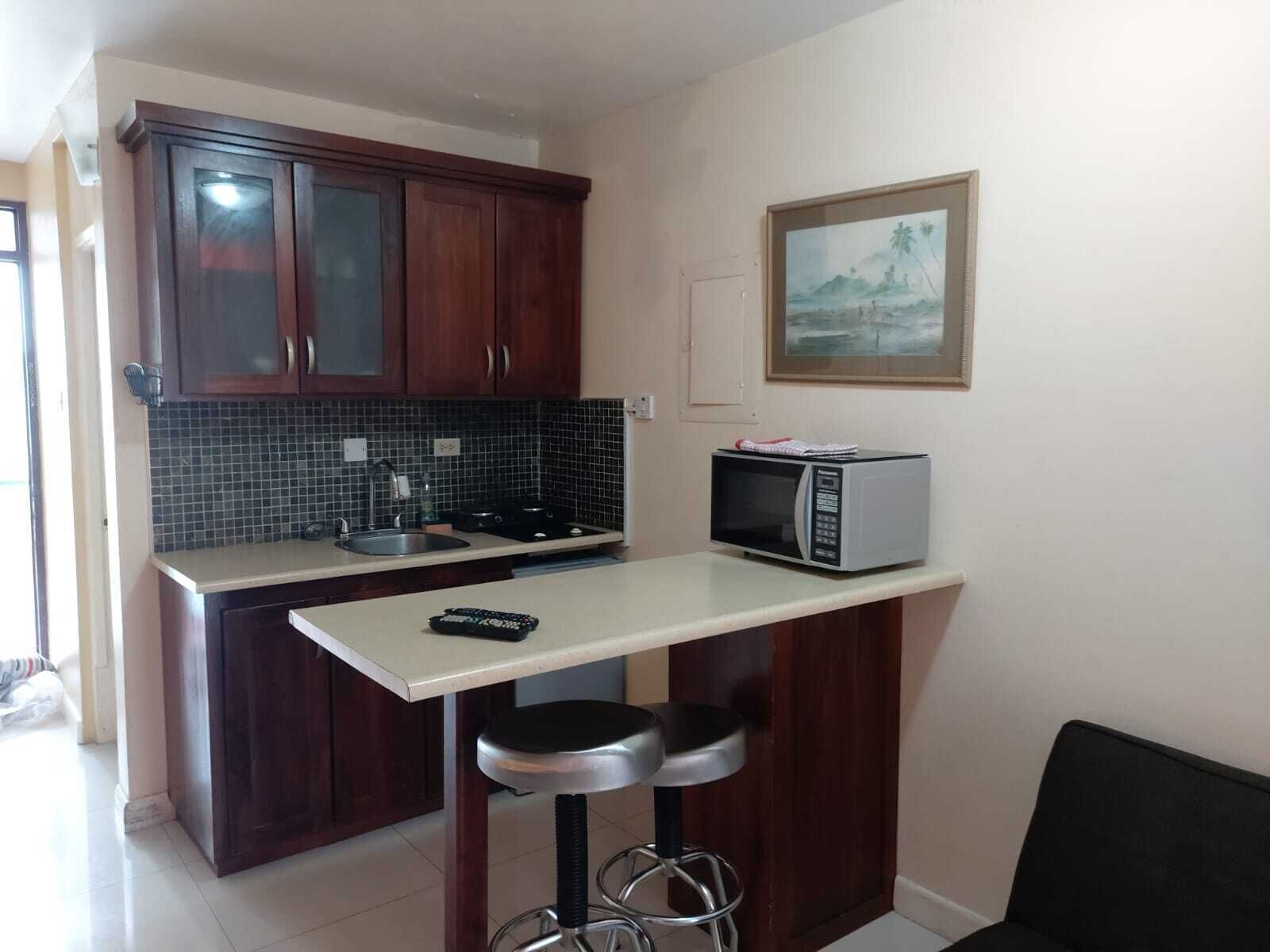 A compact kitchen with dark wooden cabinets, a tiled backsplash, a microwave, and a small bar stool.