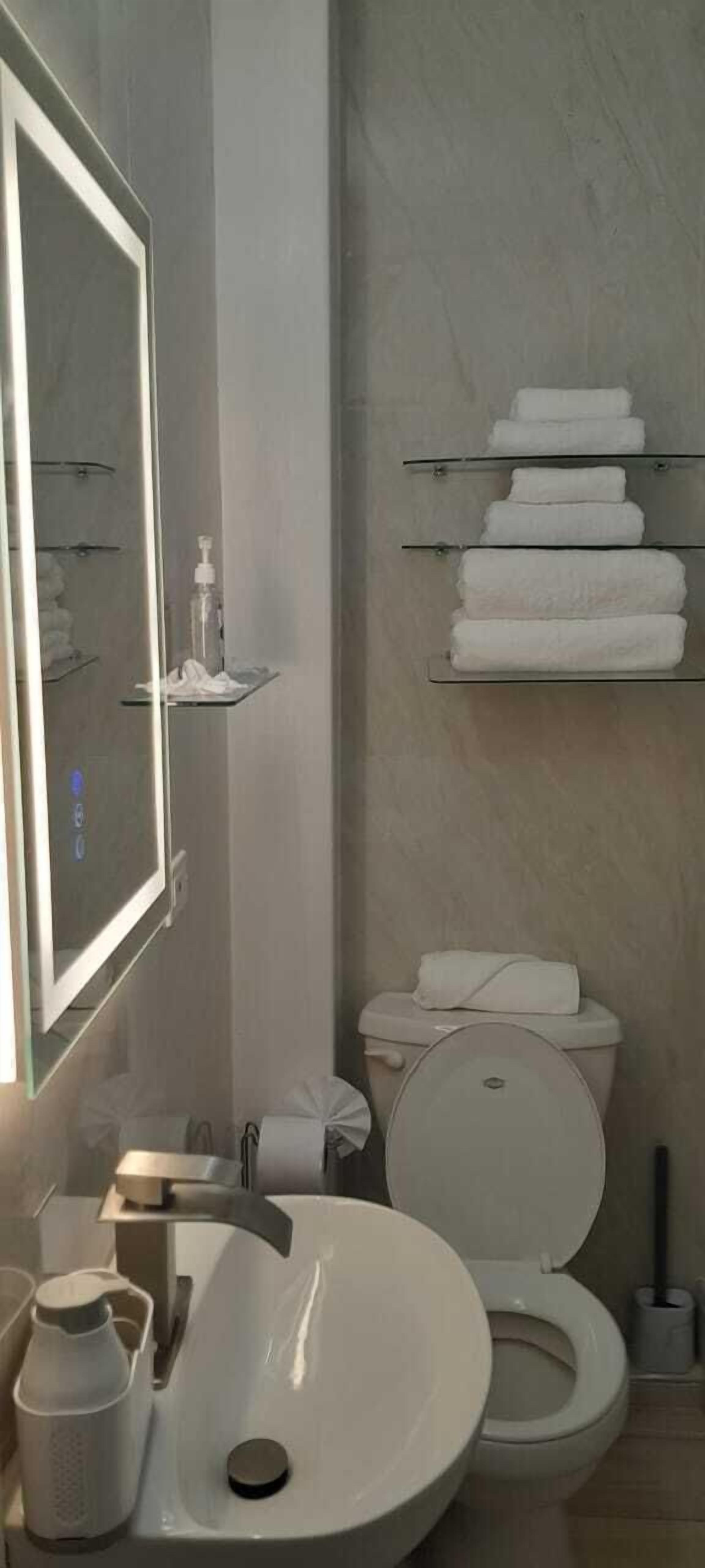 Une salle de bain avec un lavabo blanc, des toilettes et un miroir. On y trouve des étagères avec des serviettes pliées et un flacon de savon pour les mains.