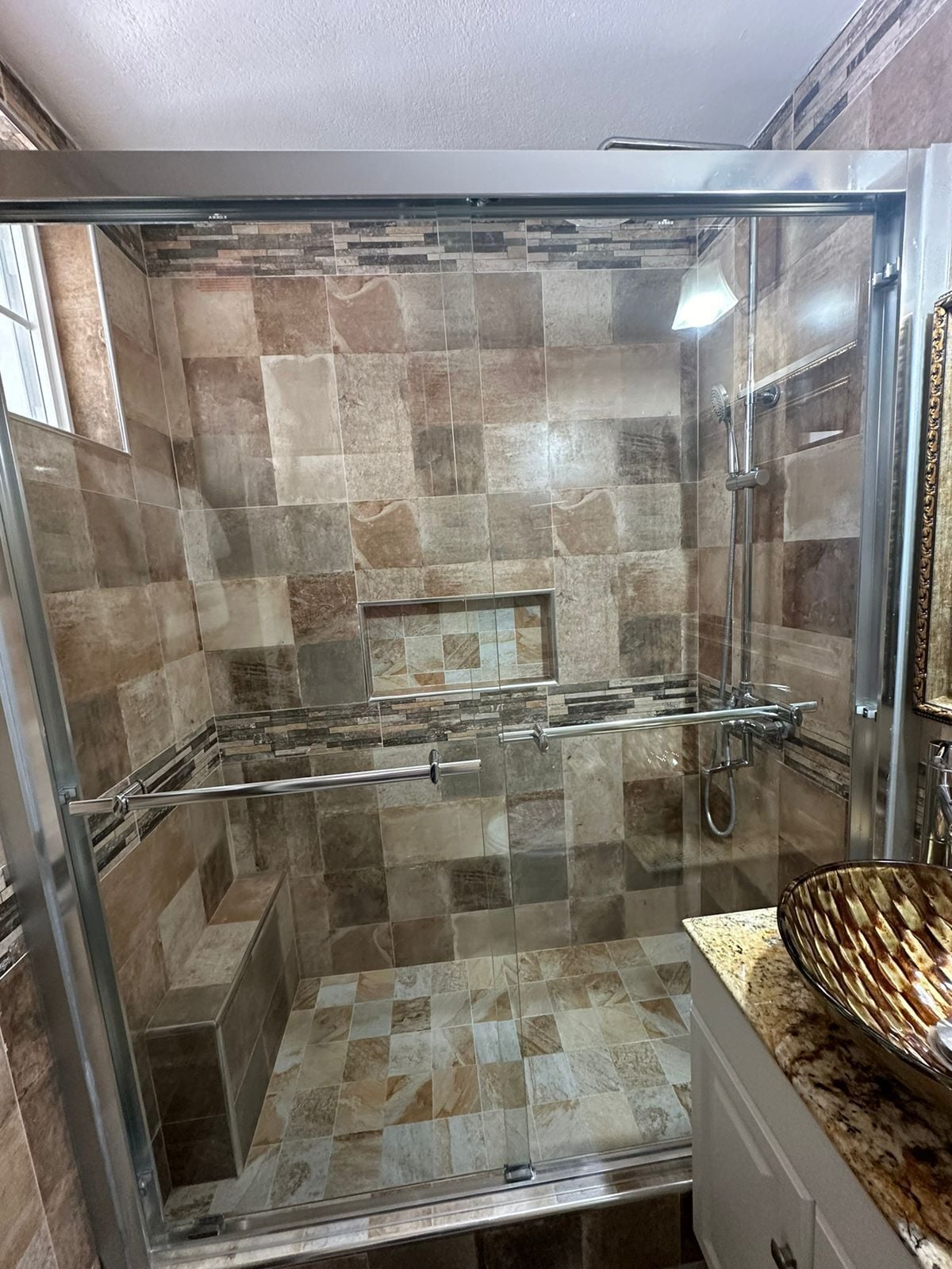 Ducha con mampara de vidrio, pared con azulejos, puerta de vidrio y encimera con un lavabo.