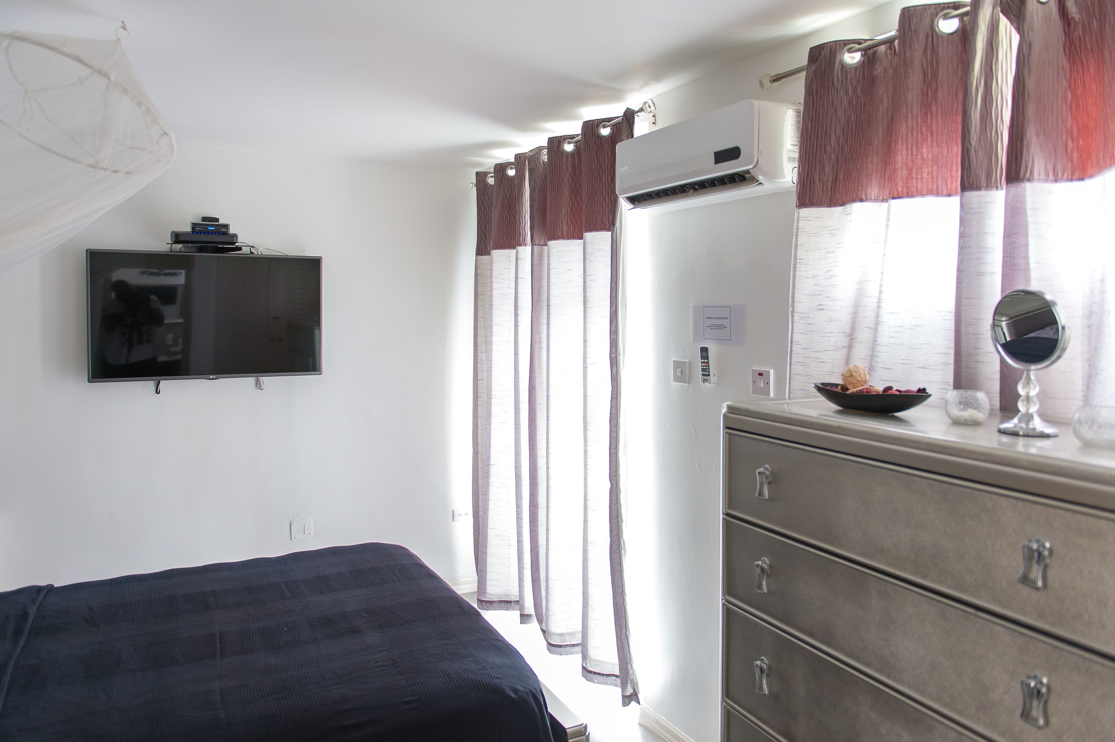 Un dormitorio con cama, cómoda, televisor montado en la pared y ventana con cortinas.