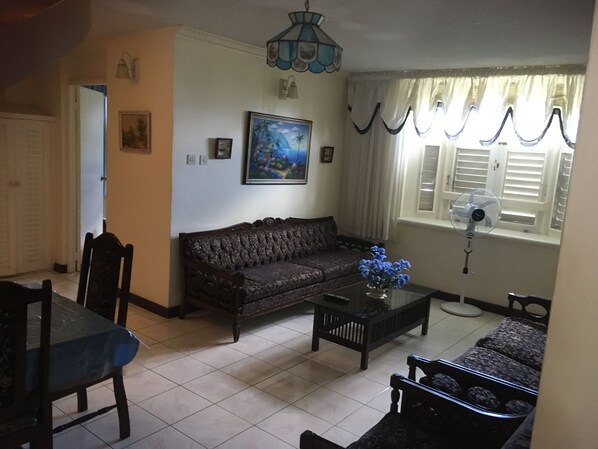A living room with a sofa, a coffee table with a vase of flowers, a dining table with chairs, a chandelier, and a window with curtains.