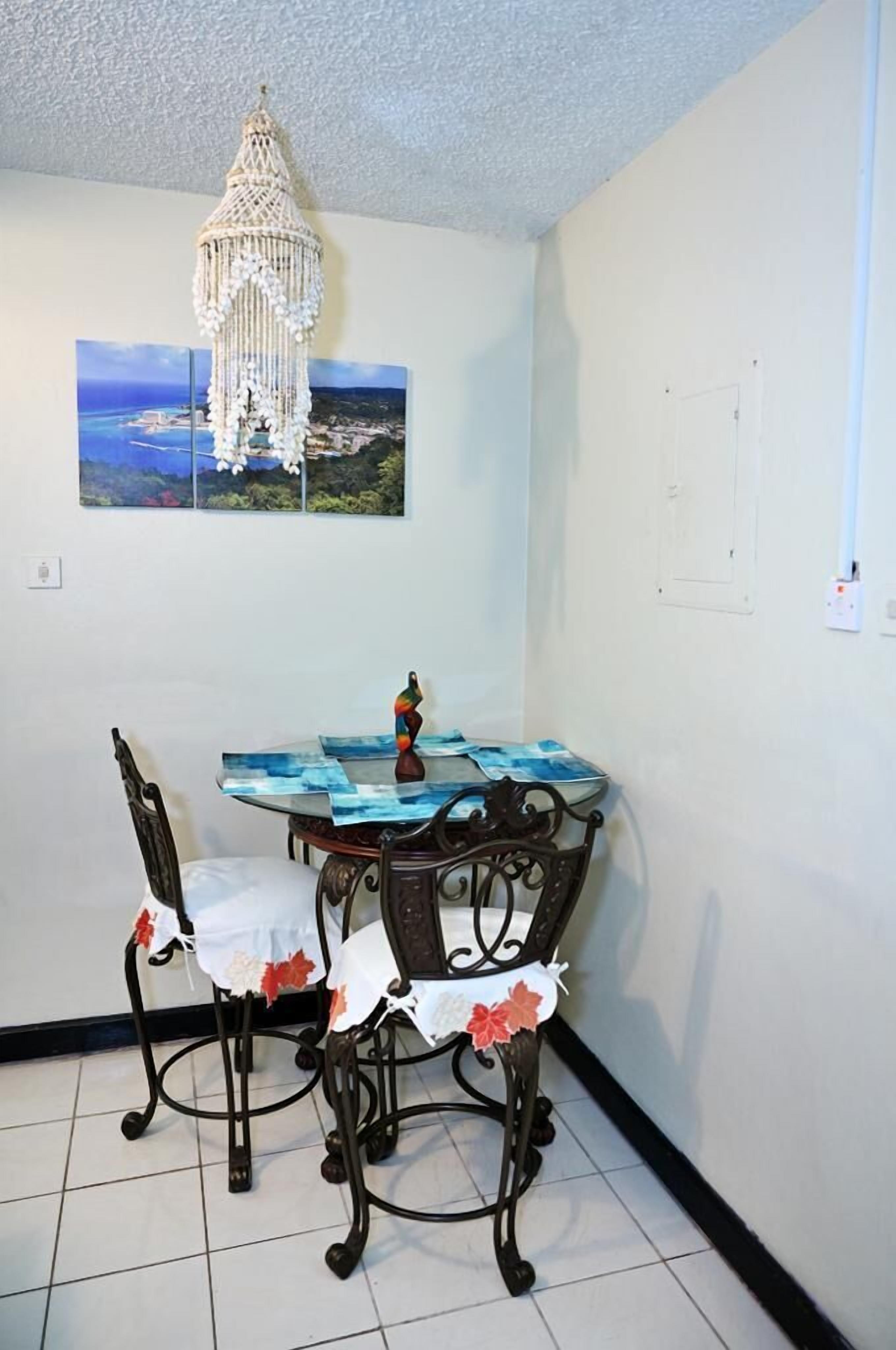 A dining area with a table set for two, a macrame chandelier, and a coastal landscape painting on the wall.