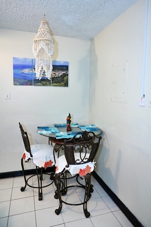 A dining area with a table set for two, a macrame chandelier, and a coastal landscape painting on the wall.