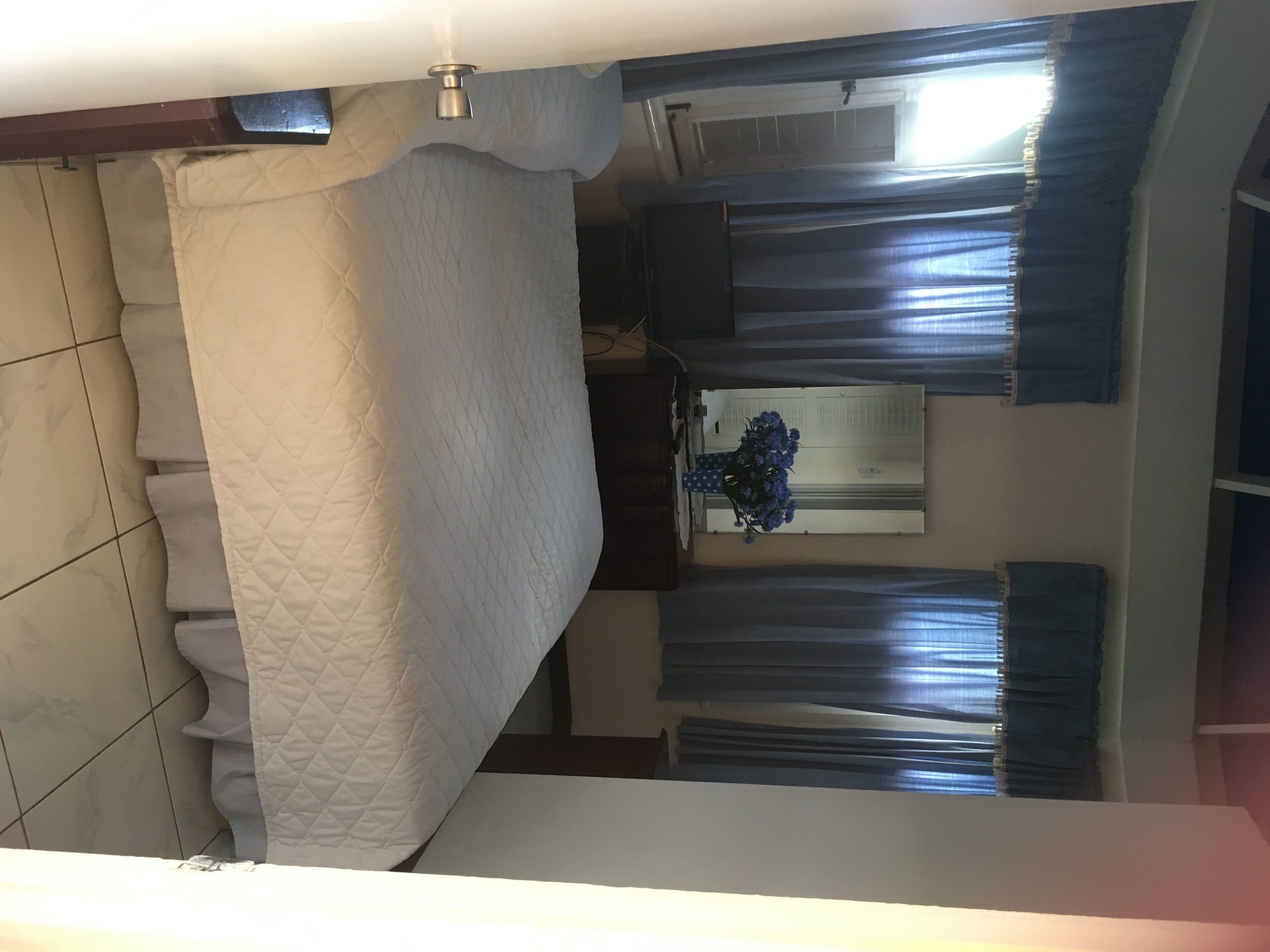 A room with a bed, a window with curtains, a vase with flowers, and a tiled floor.