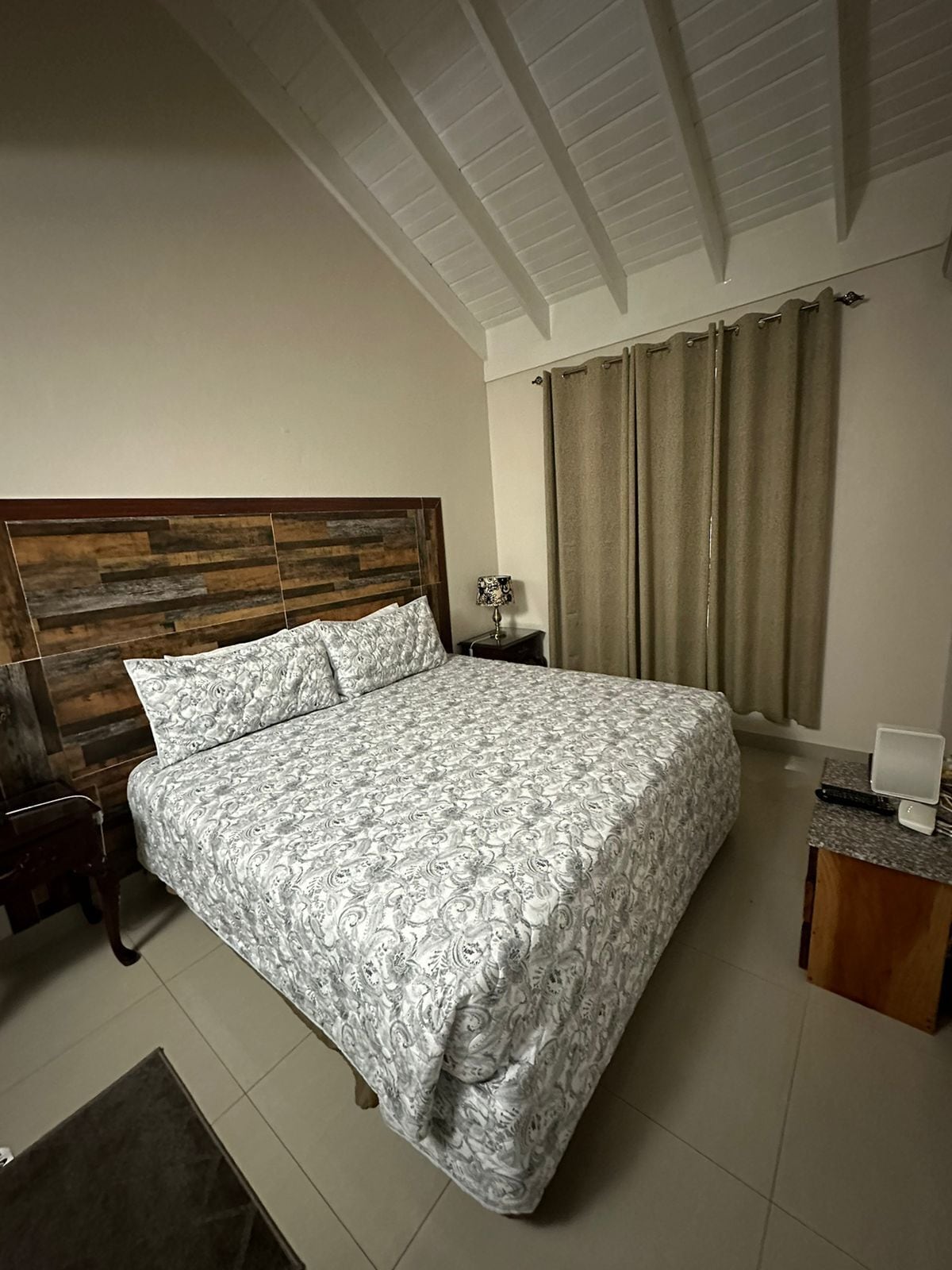 Un dormitorio con cabecera de madera, colcha estampada, mesita de noche con lámpara y escritorio con computadora.