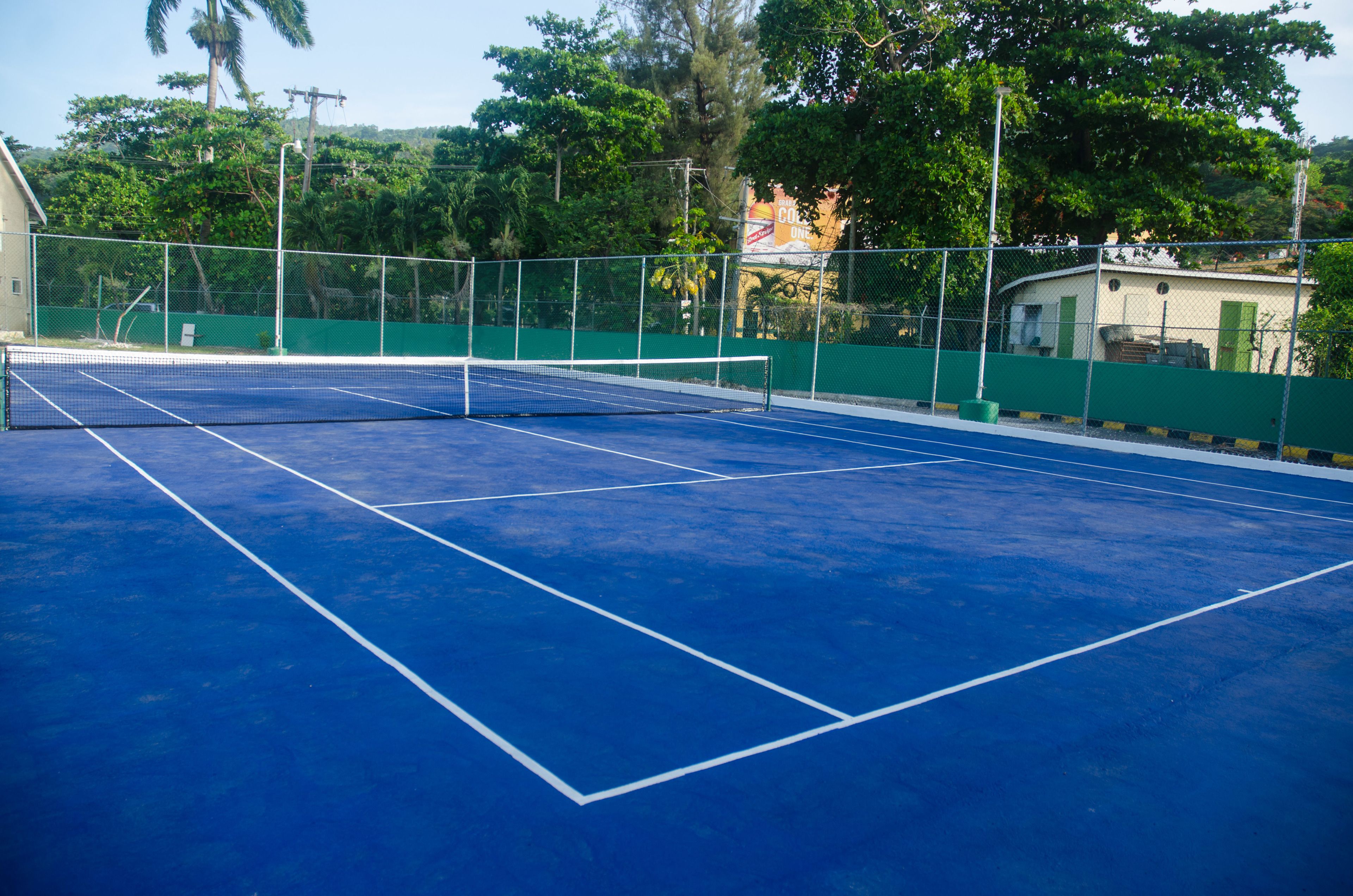 Una cancha de tenis al aire libre con una red, rodeada por una cerca verde y árboles.