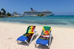 Two beach chairs with colorful covers, a cruise ship in the background, and clear blue water.