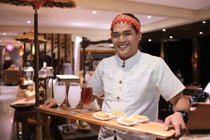 Uma pessoa de avental de chef segurando uma bandeja com sobremesas e bebidas em ambiente interno bem iluminado.