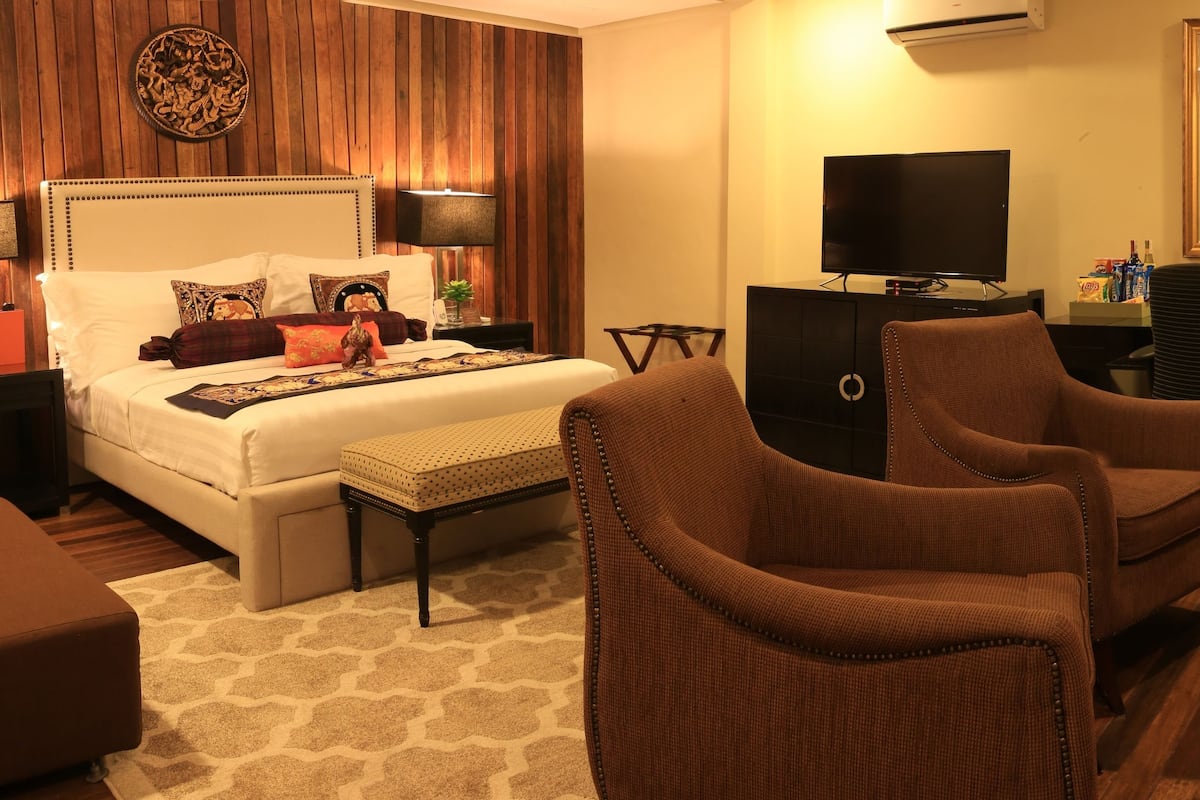 A modern hotel room with a large bed, two bedside tables, a TV, a desk, and two brown armchairs.