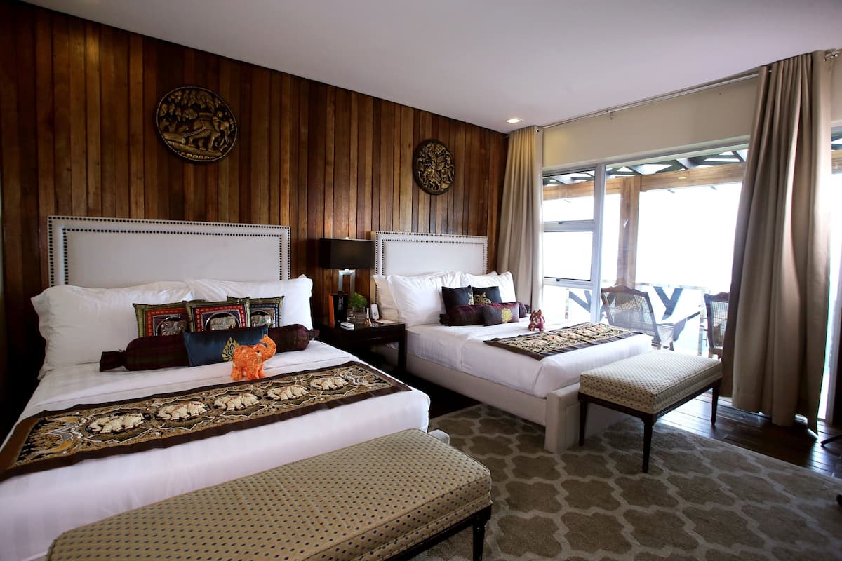 A hotel room with two beds, a wooden wall panel, a large window with curtains, and a view of the outside.