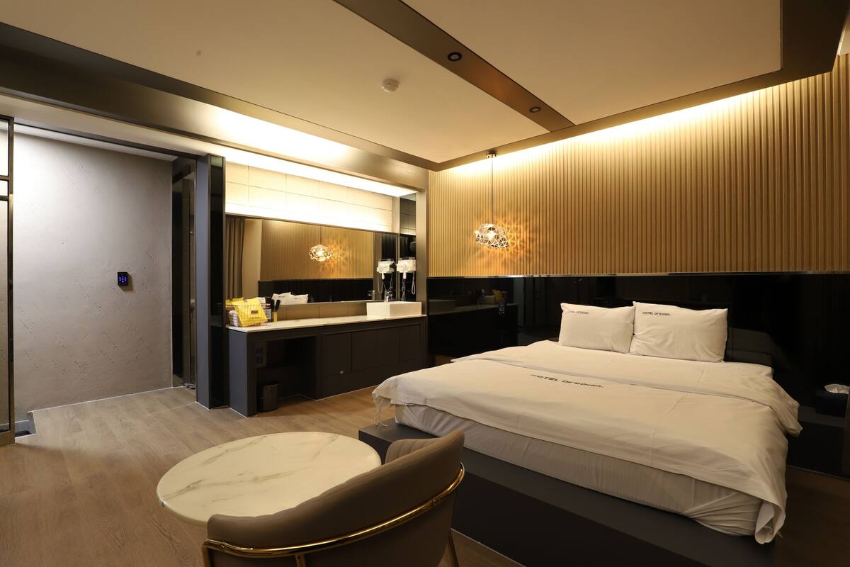 A modern hotel room with a large bed, a separate seating area, a bathroom with a mirror, and a minibar.