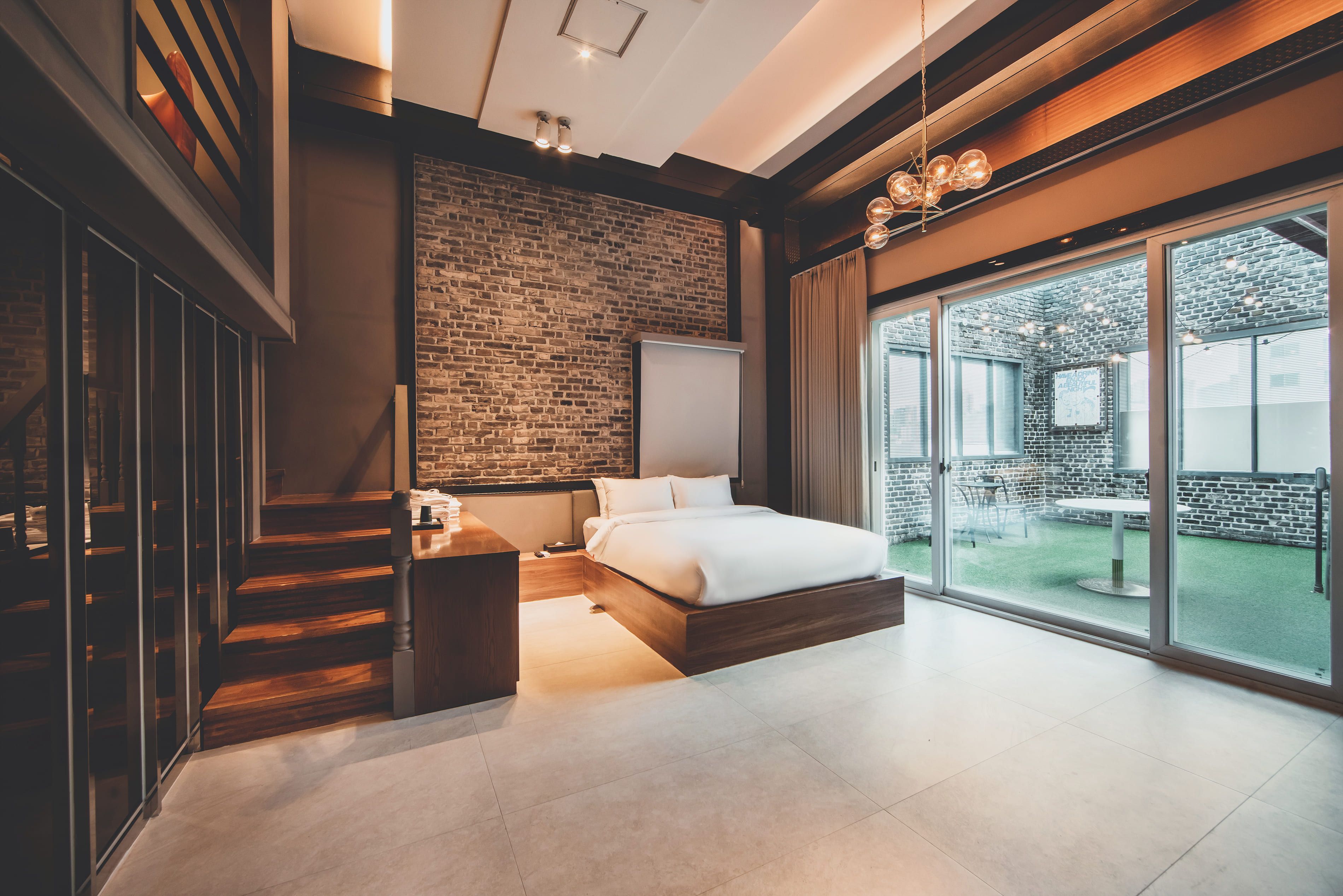 Un dormitorio moderno con pared de ladrillo, una cama grande, una escalera de madera y un ventanal corredero que da a un área exterior.