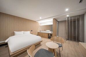 A modern hotel room with a large bed, a small desk, a chair, and a shower area.