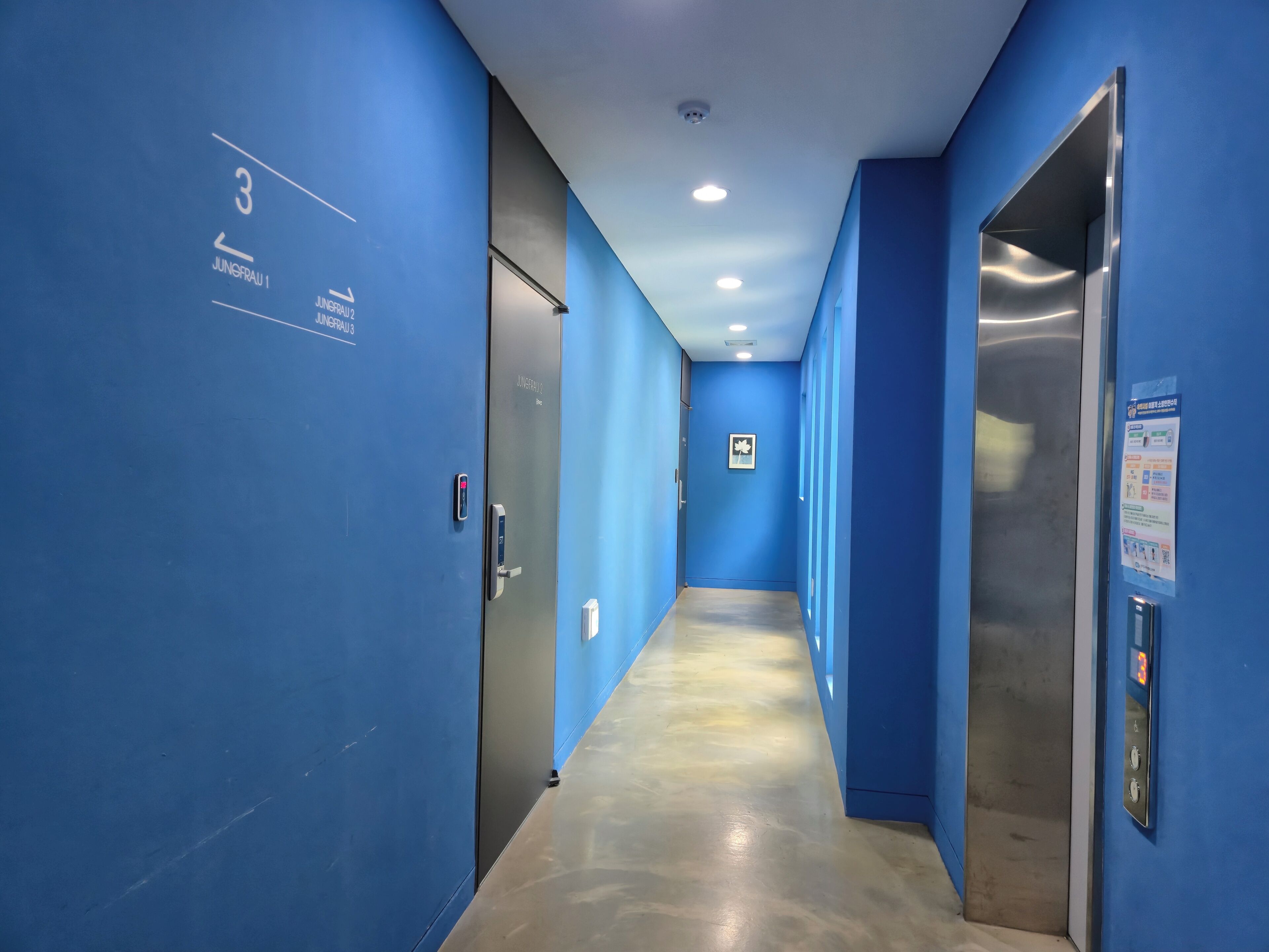 Un couloir aux murs bleus, avec des portes numérotées et un ascenseur.