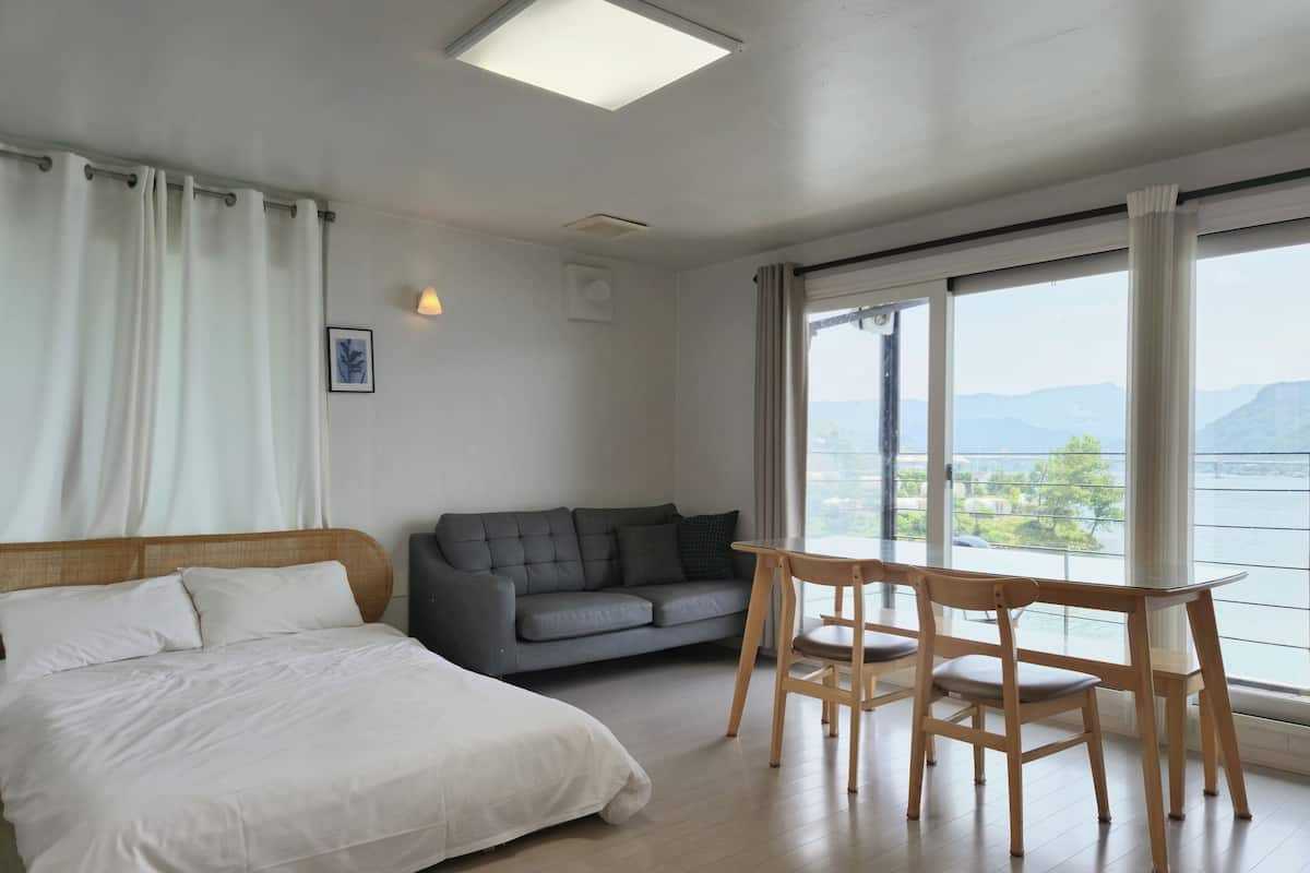 A bedroom with a bed, a grey sofa, a wooden table, and a large window with a view of a body of water and mountains.