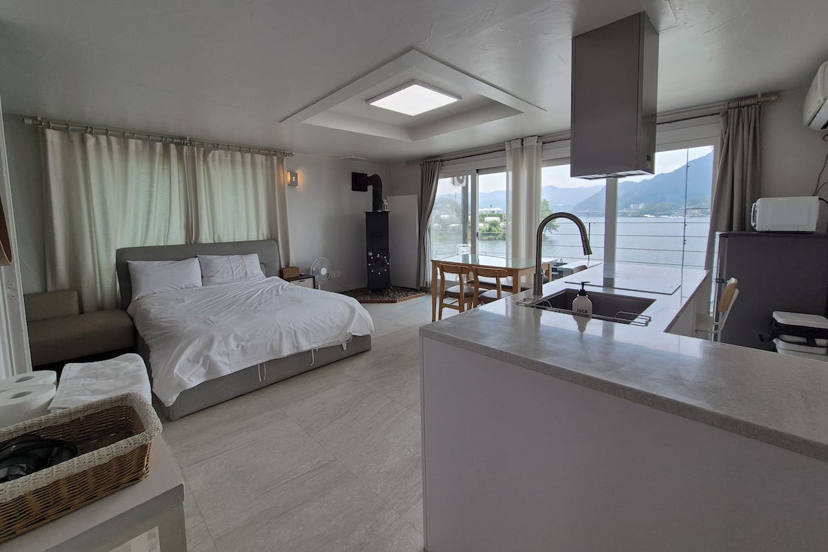 A modern hotel room with a bed, a small dining area, a kitchen with a sink, and a large window offering a view of the sea and mountains.