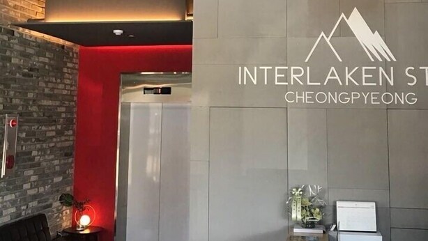 Un intérieur moderne avec un comptoir d’accueil, une enseigne fixée au mur indiquant « Interlaken St. Cheongpyeong », et un mur d’accent rouge.