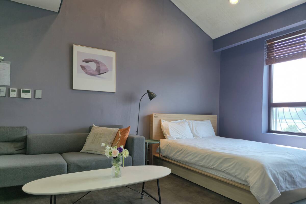 A modern bedroom with a grey sofa, a white bedside table with a vase of flowers, a bed with white bedding, and a large window with blinds.
