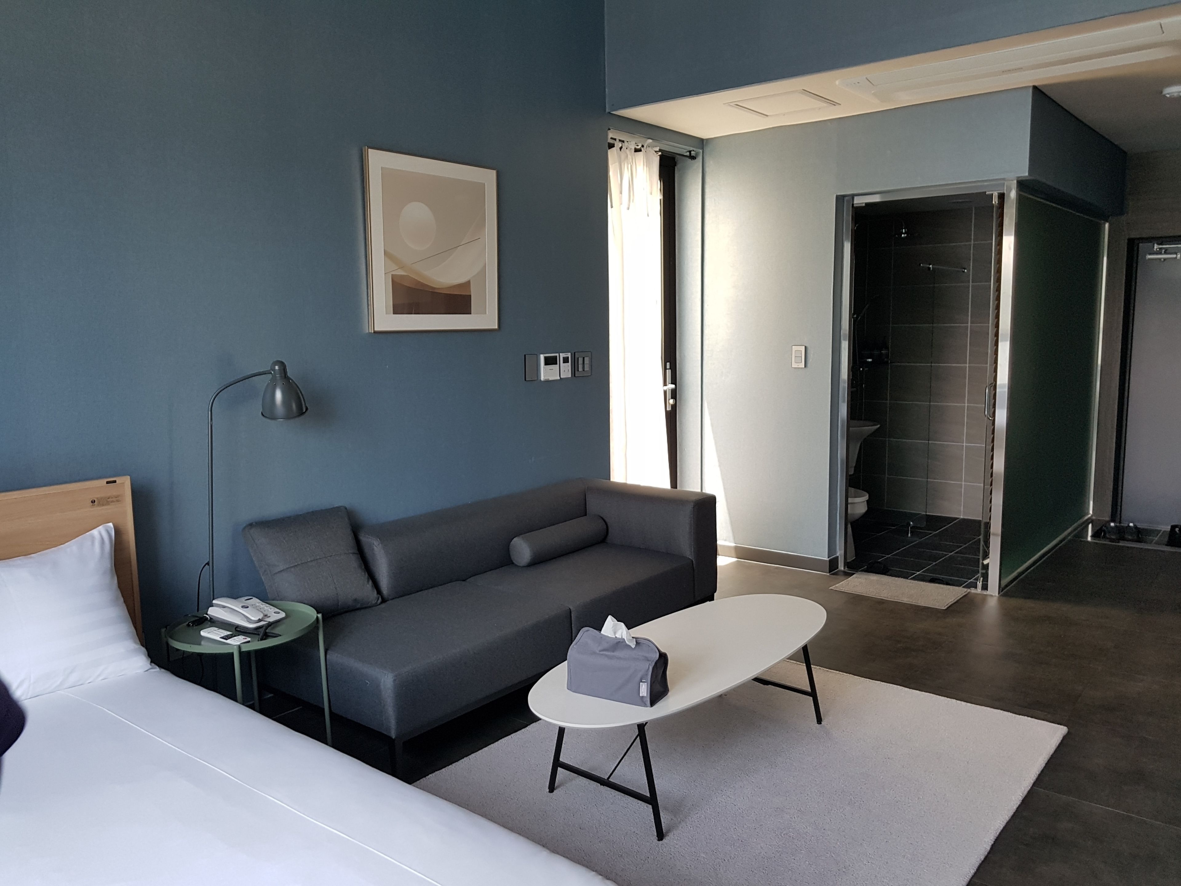 A modern hotel room with a grey sofa, a small round table, a bed, a bedside lamp, a framed artwork on the wall, and a bathroom visible through an open door.