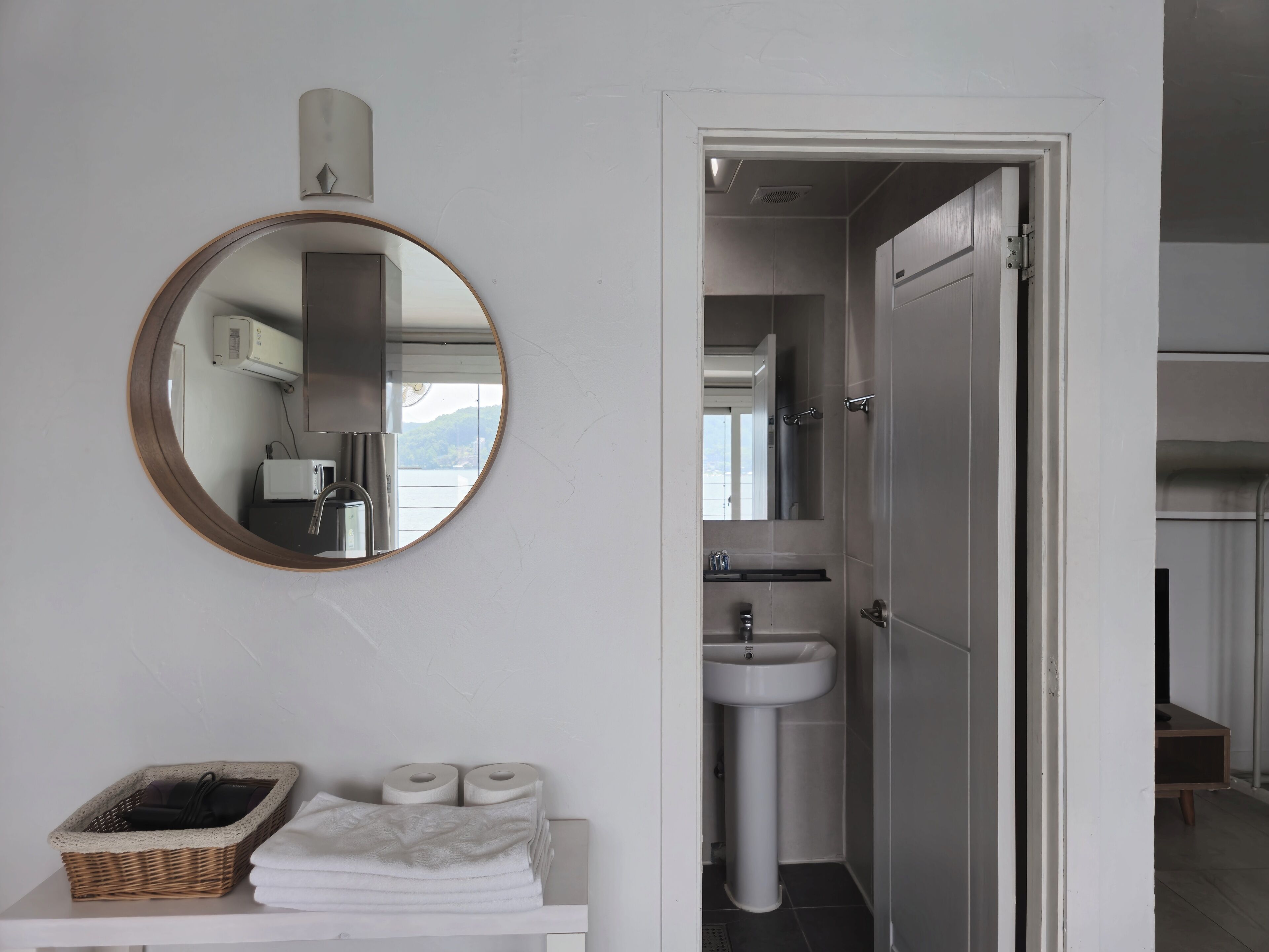 A bathroom with a round mirror, a sink, and a toilet. There is a basket with towels and toilet paper on a shelf.