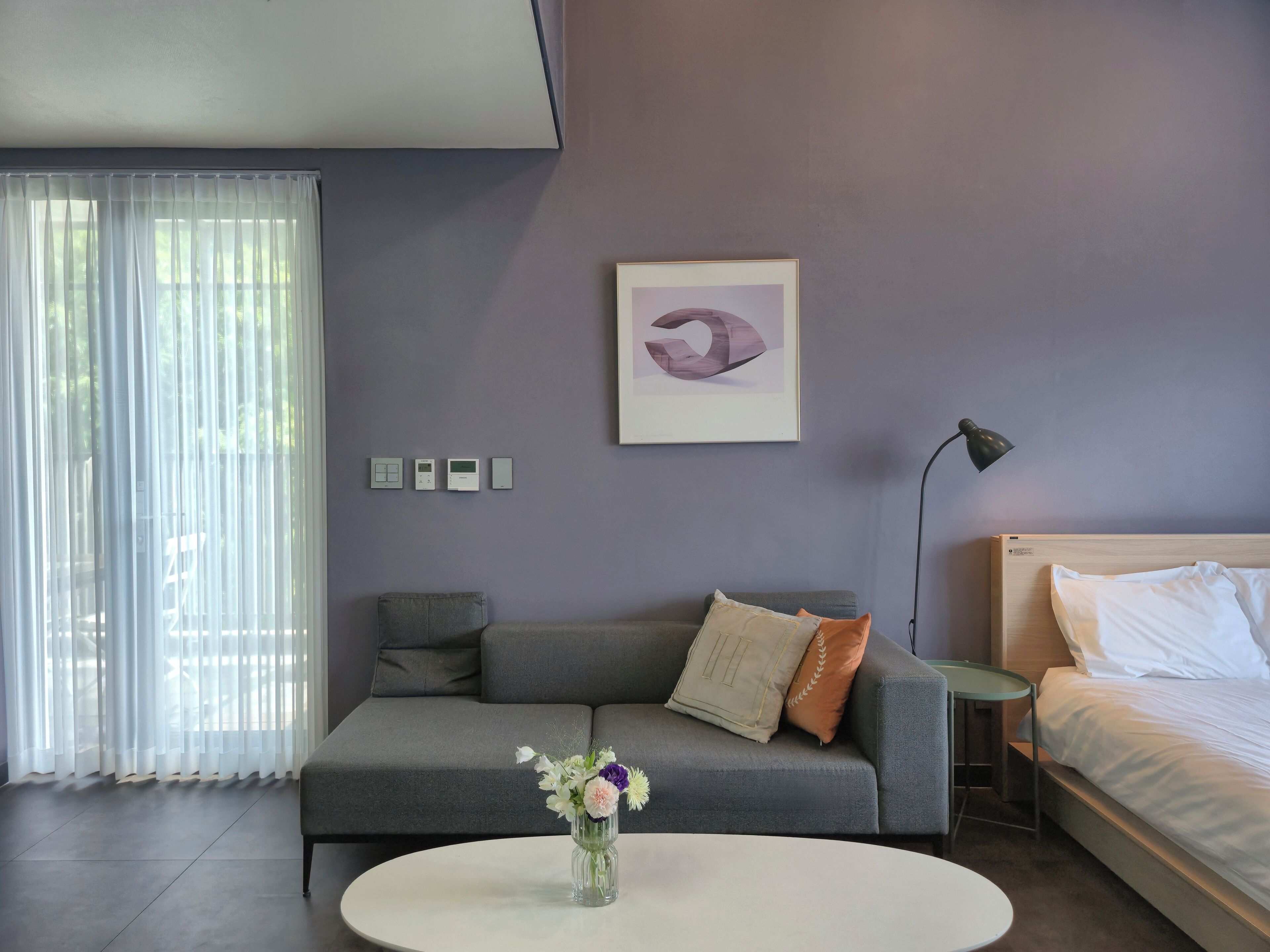 Une chambre d’hôtel moderne avec un canapé gris, une petite table ronde ornée d’un vase de fleurs, un lit avec du linge de maison blanc et une œuvre d’art abstraite accrochée au mur.