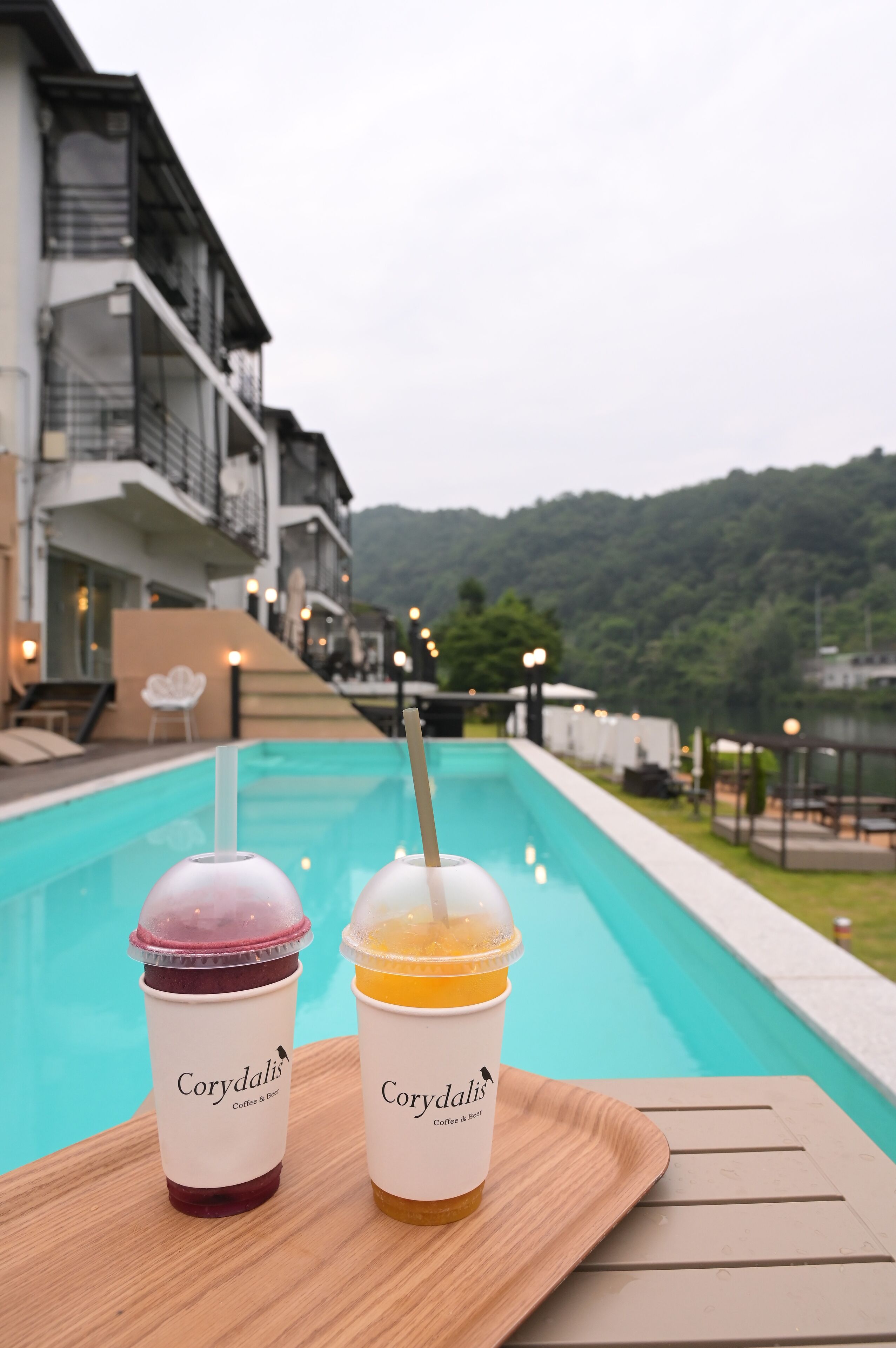 Dos bebidas heladas sobre una mesa de madera junto a una piscina, con vista a un edificio y colinas al fondo.