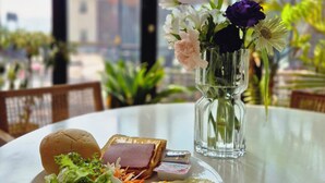 Une table à manger avec une assiette de nourriture, un vase rempli de fleurs et une vue sur les bâtiments à travers de grandes fenêtres.