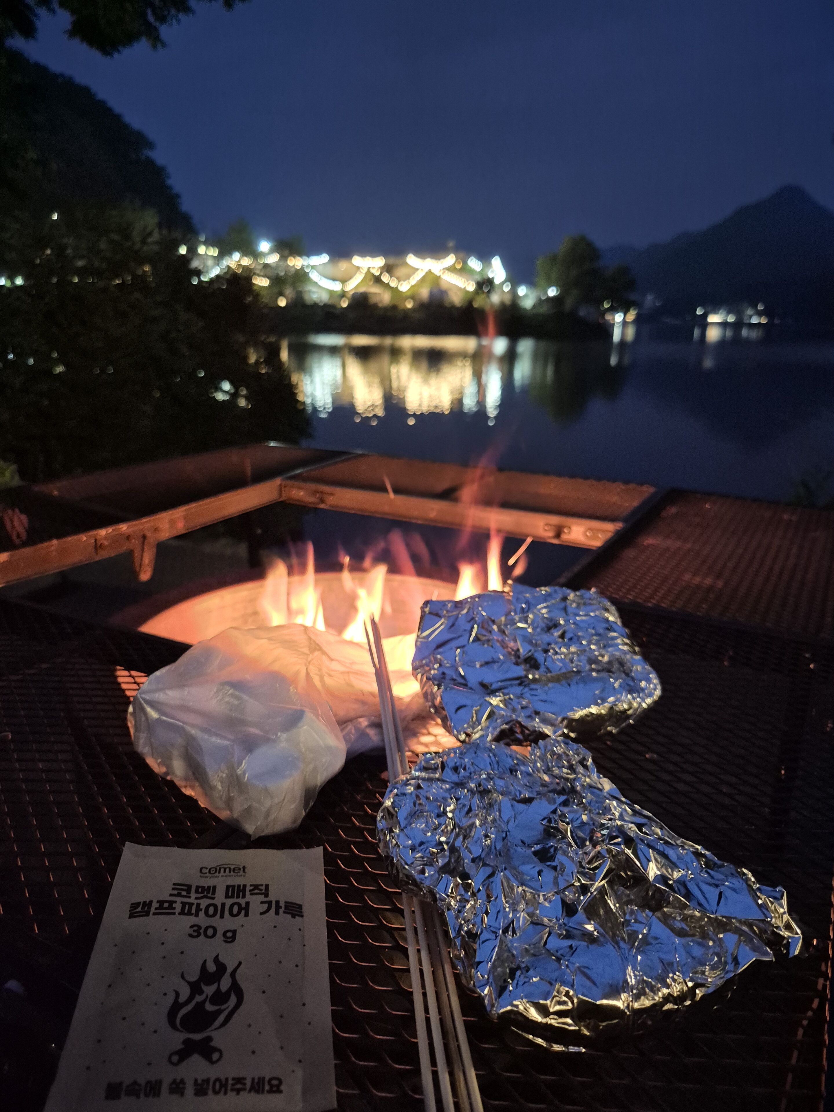 A grill with foil-wrapped food items and a menu on a table by a lake at night.