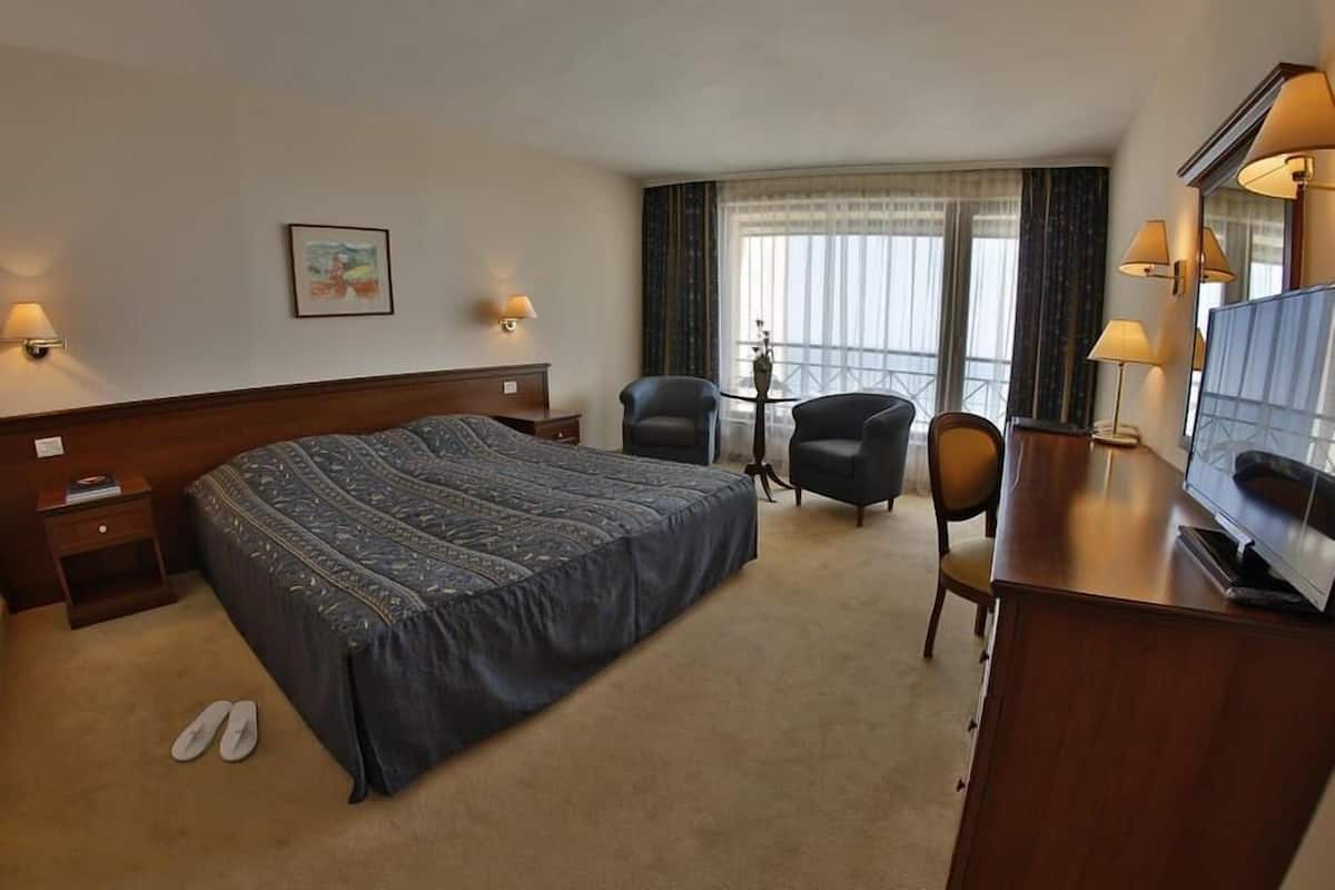A hotel room with a large bed, two armchairs, a desk with a television, and a window with curtains.