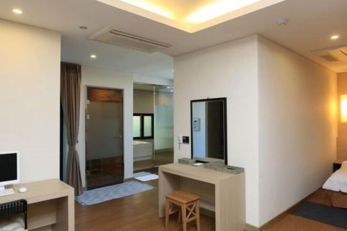 A modern hotel room with a desk, a small stool, a bed, and a mirror.