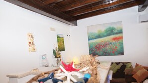 Une pièce avec un plafond en bois, un mur blanc orné d’œuvres d’art, un panier en osier contenant divers objets et un canapé en osier avec des coussins.